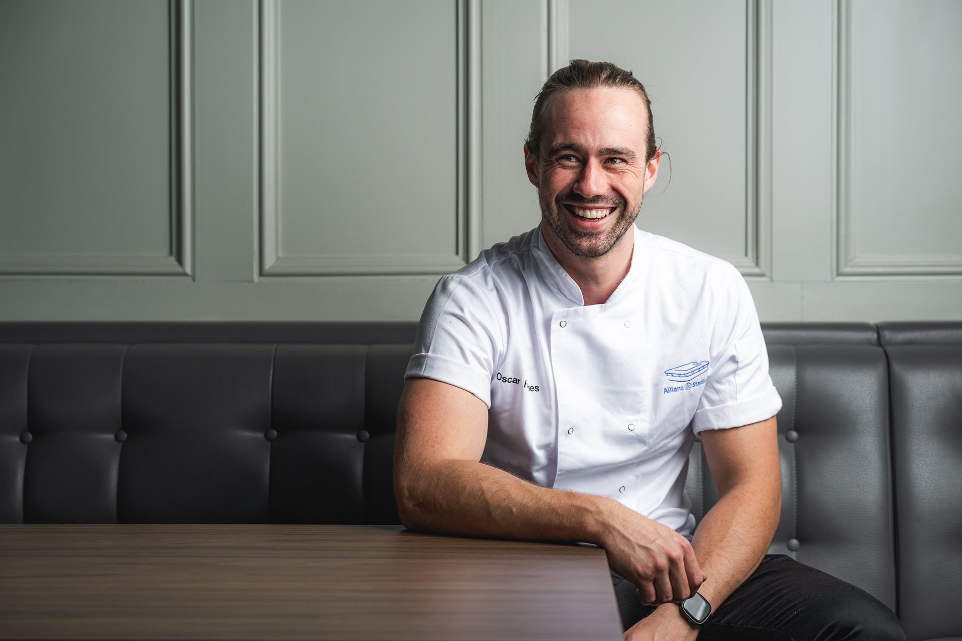 A smiling chef leaning forwards beside a table, seated against a grey panelled wall.