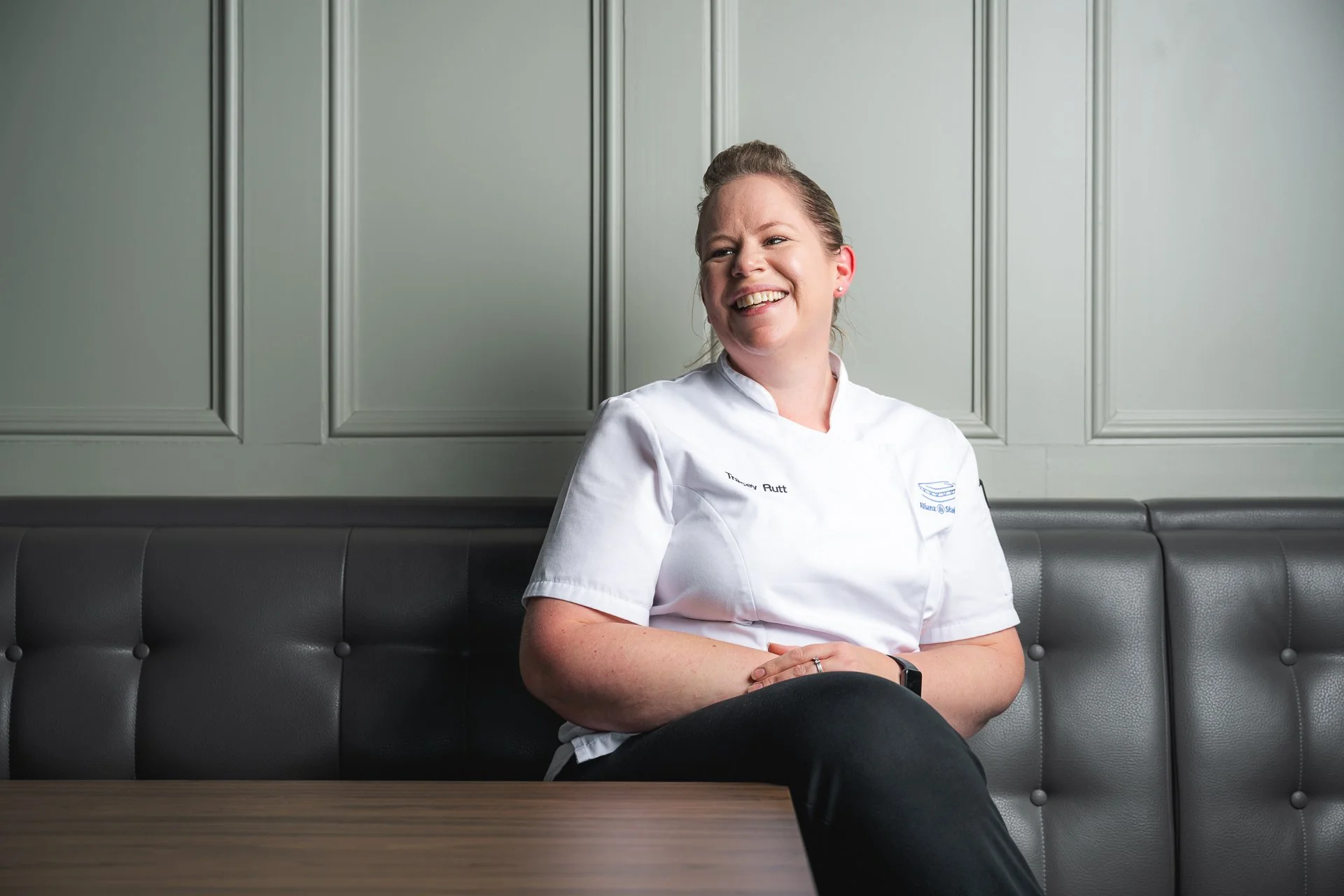 A smiling chef seated on a dark upholstered bench in front of a grey panelled wall.