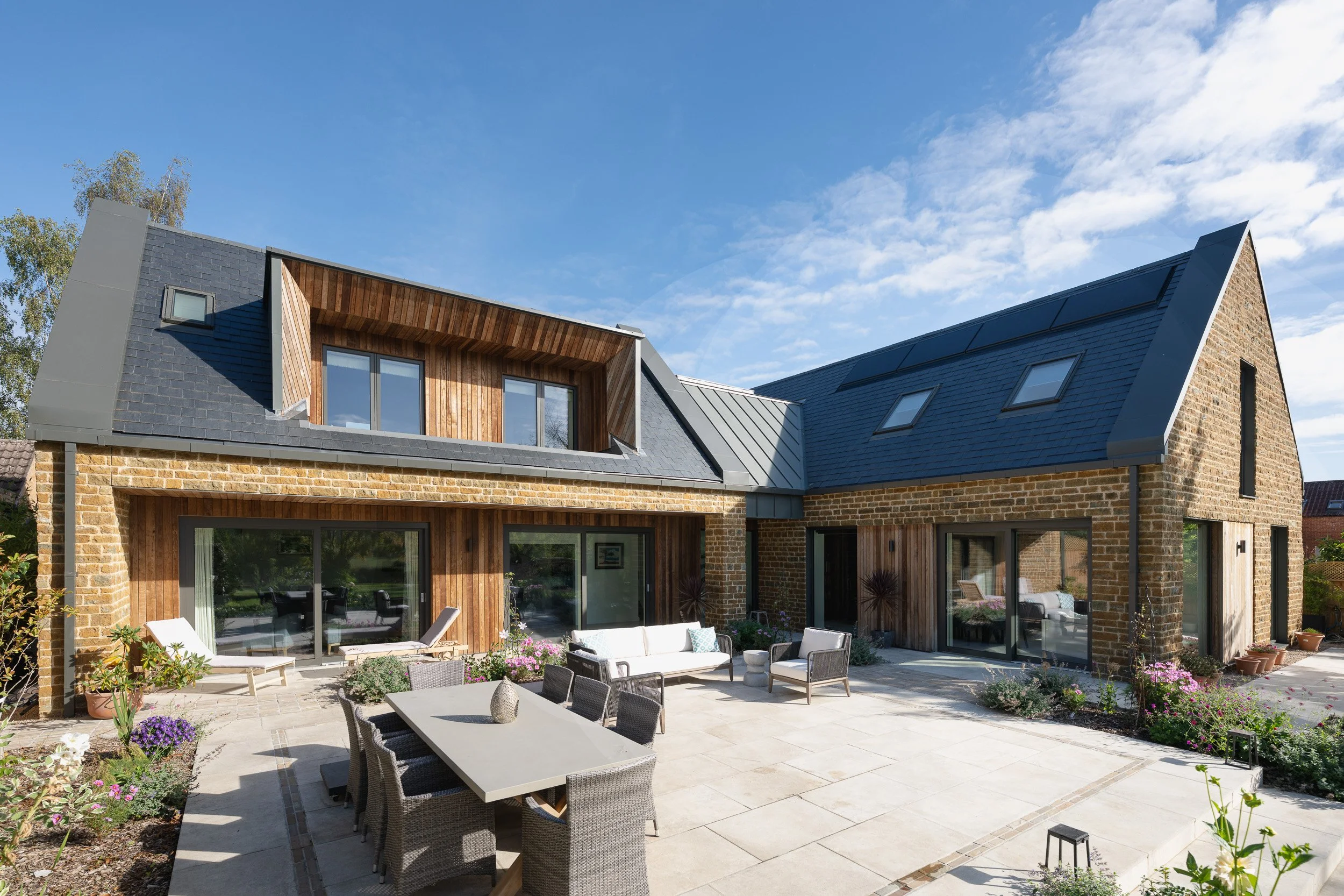 Wide view of a paved terrace with outdoor furniture, bordered by the stone and timber house.