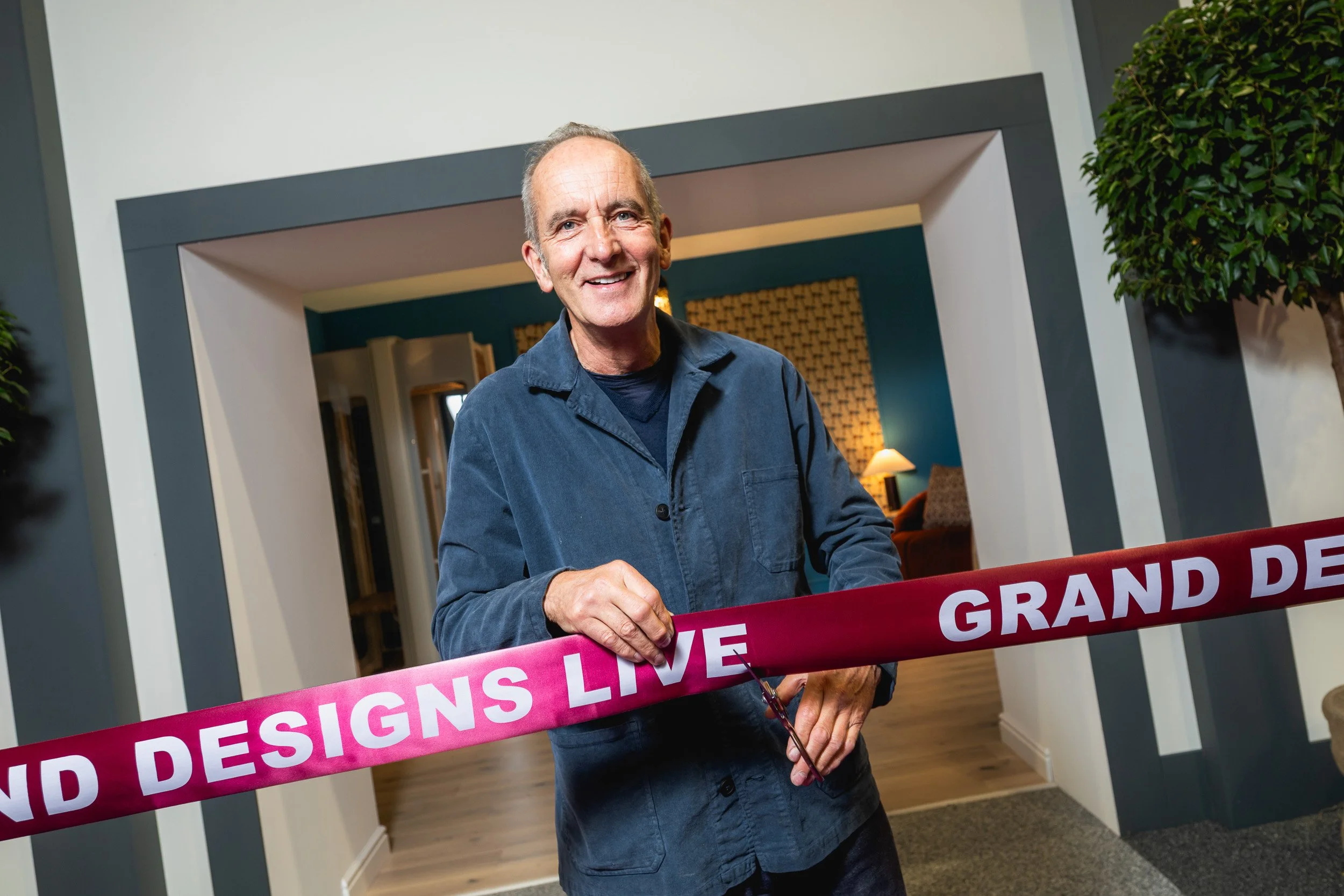 Kevin McCloud holding scissors beside a Grand Designs Live ribbon at the NEC.