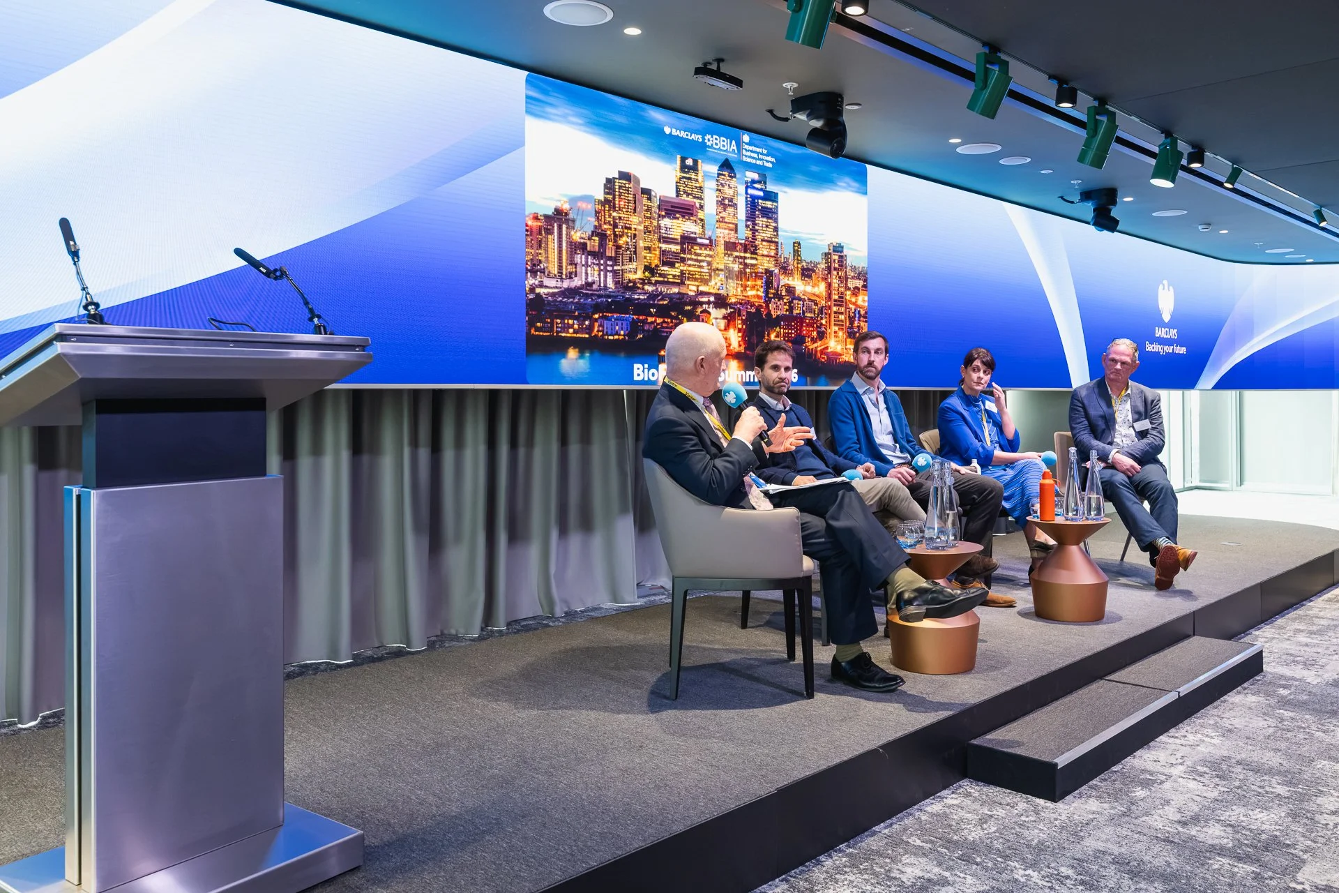 Panel discussion at the UK BioFinance Summit 2026, with five people seated on stage beneath a wraparound screen at Barclays HQ in Canary Wharf.