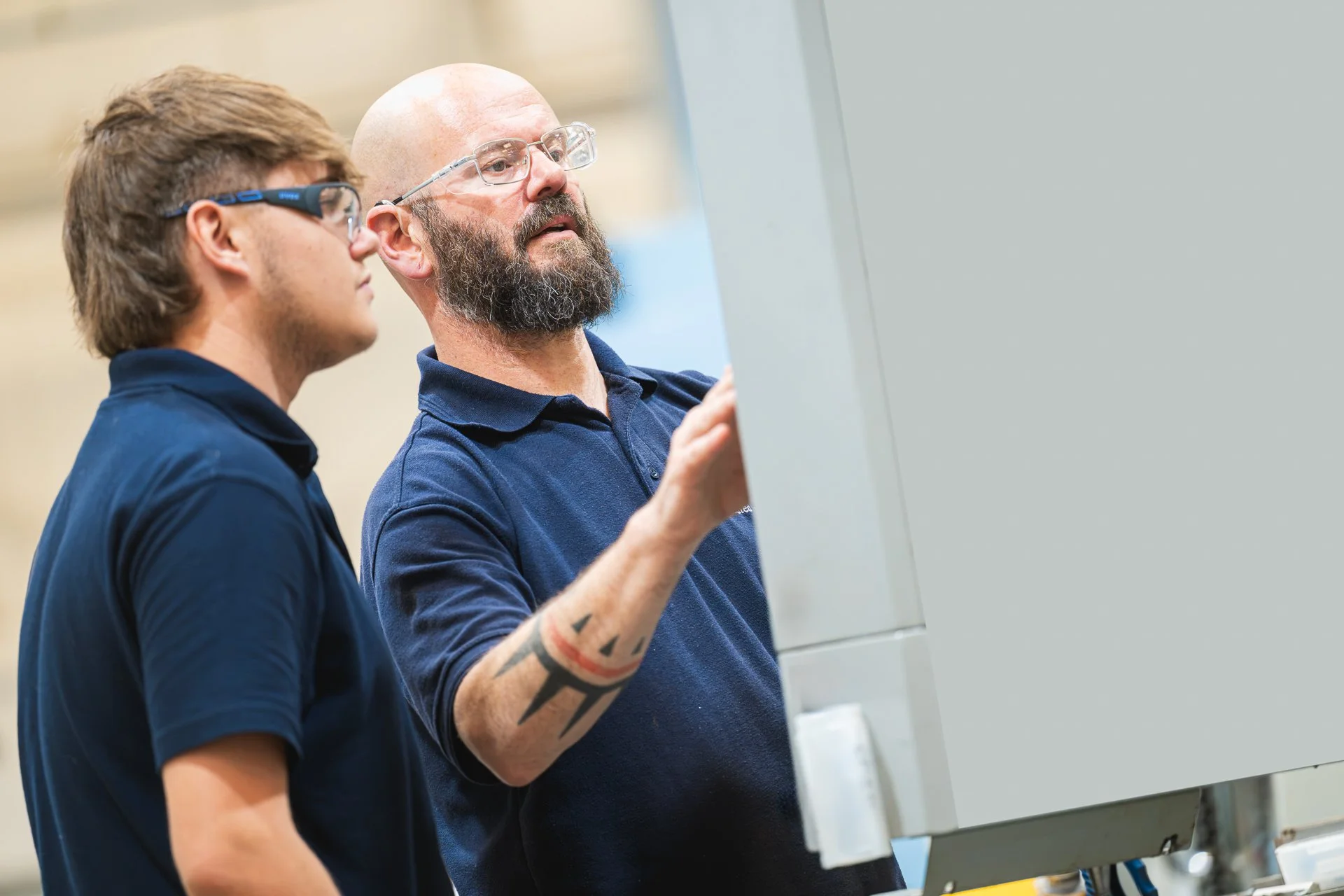 Two colleagues in navy polo shirts looking towards a machine control area.