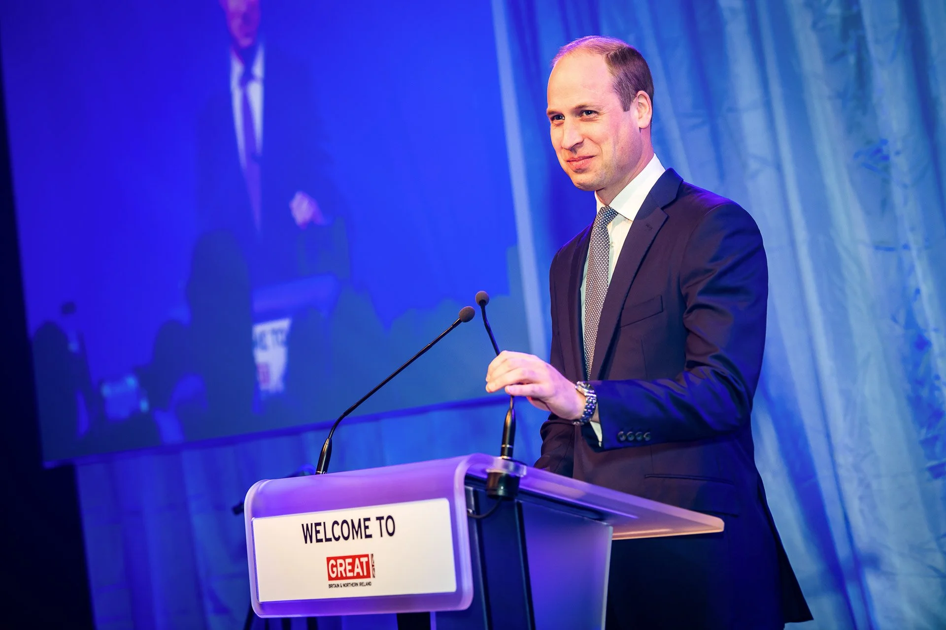 Prince William delivering a presentation from a lectern on a blue-lit stage.