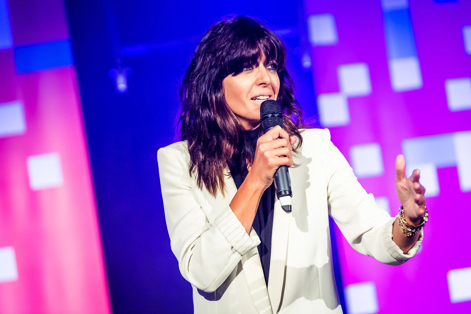 Claudia Winkleman in a white jacket addressing the audience with a handheld microphone.