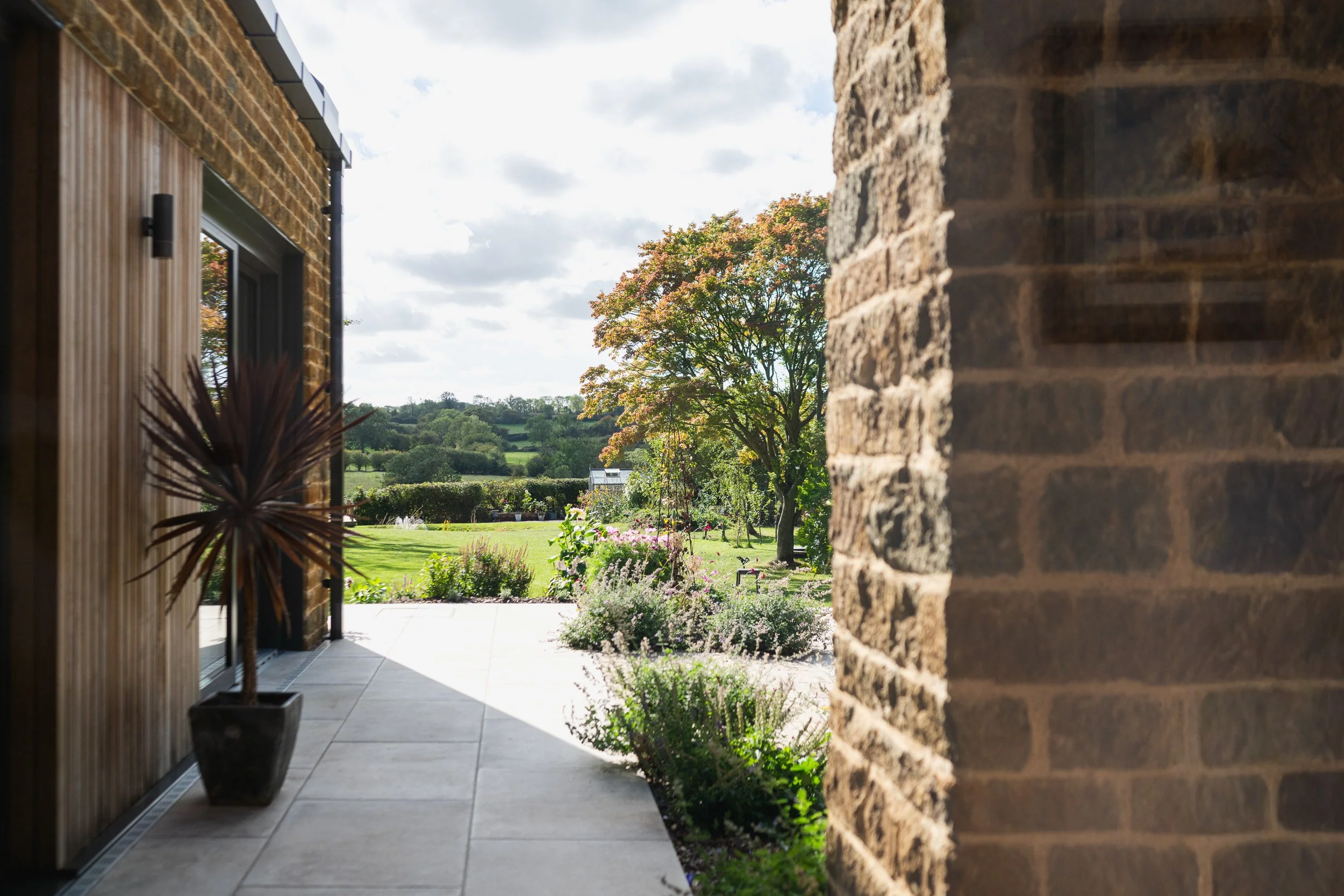 View along a paved passage between stone and timber walls towards a planted garden and countryside.