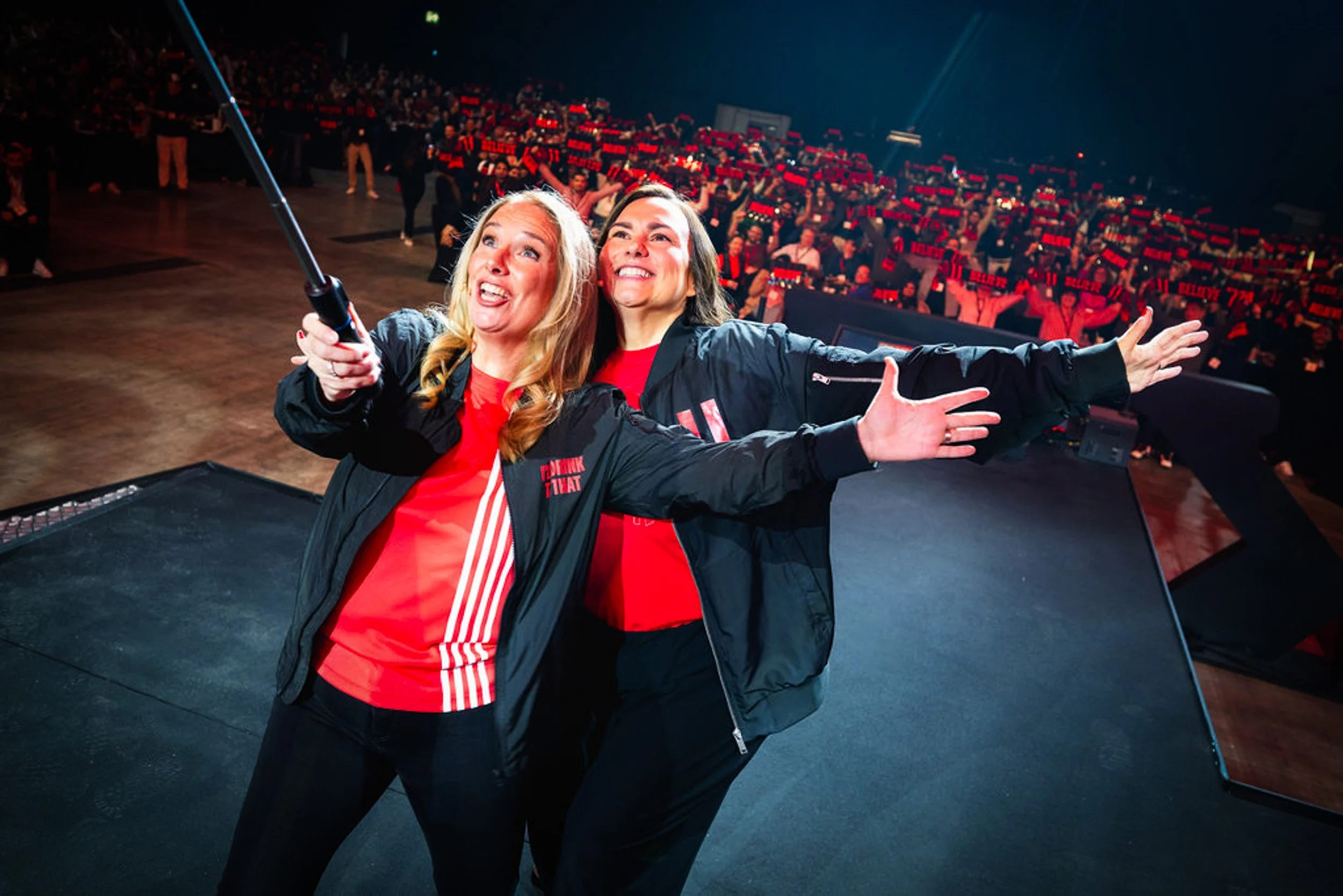 Two people taking a selfie on stage, with KFC delegates holding BELIEVE scarves behind them.