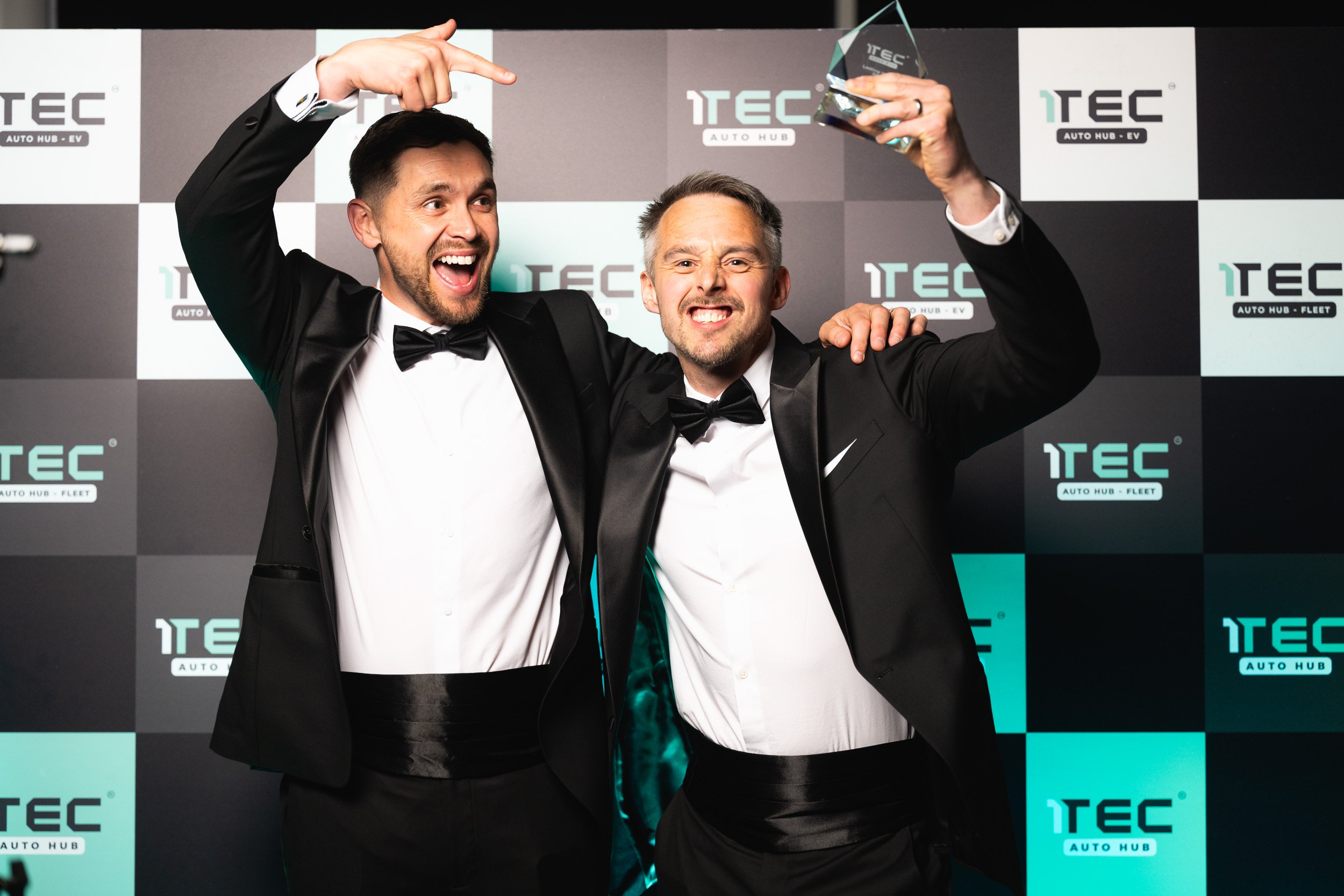 Two guests in dinner jackets celebrating with a raised trophy in front of a 1TEC backdrop.