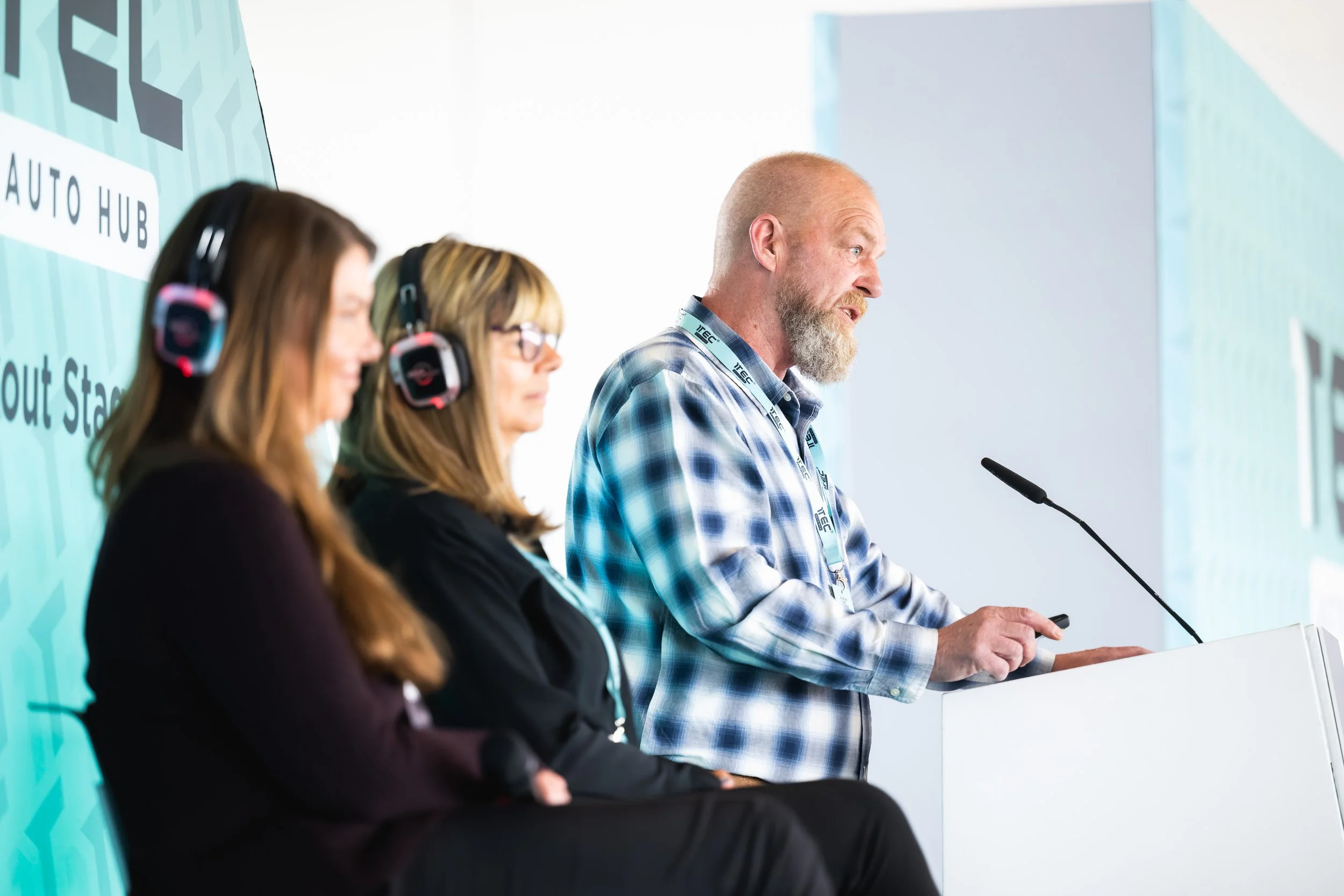 Speaker at a lectern beside two seated participants wearing headphones.