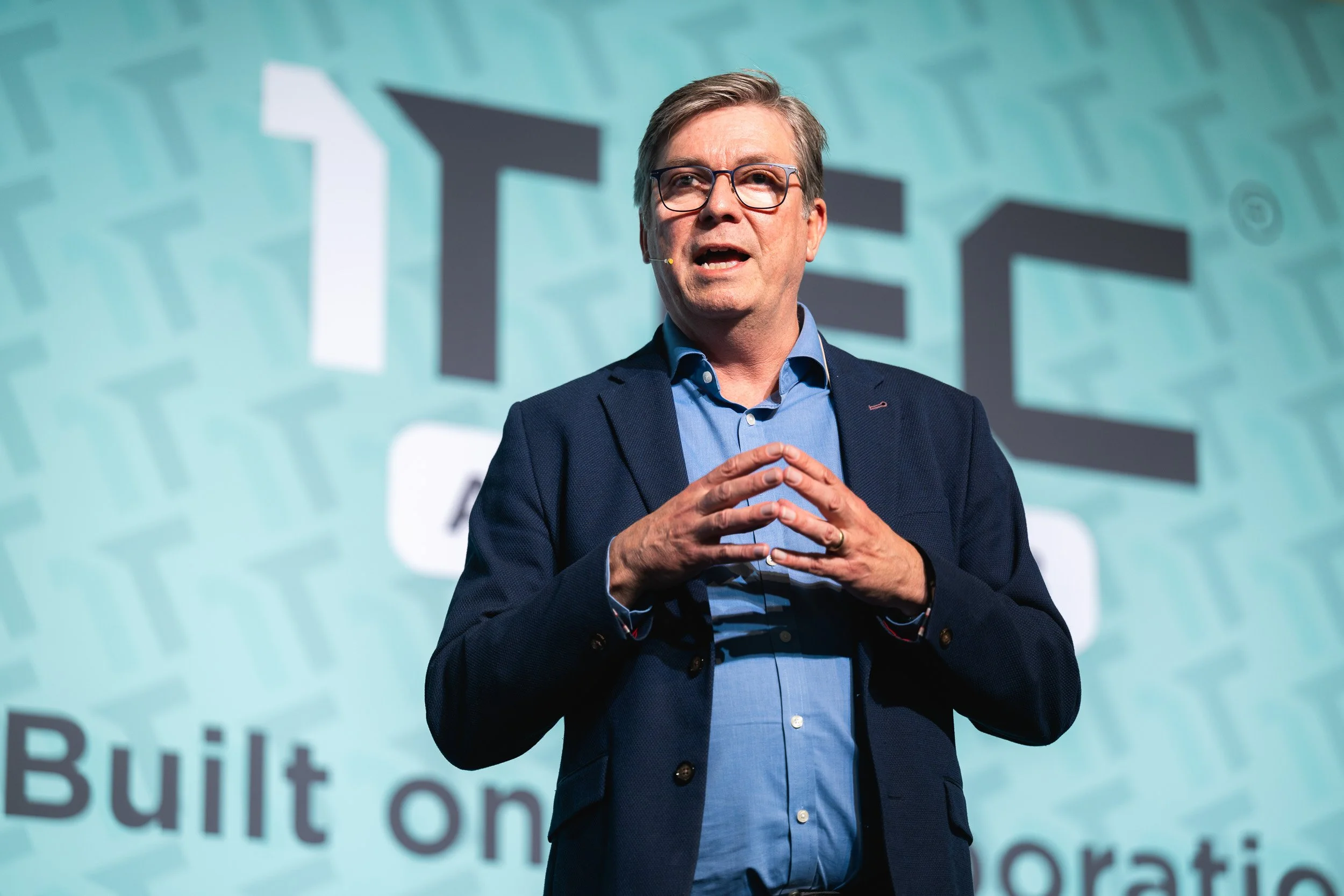 Speaker addressing the 1TEC conference in front of a branded turquoise backdrop.