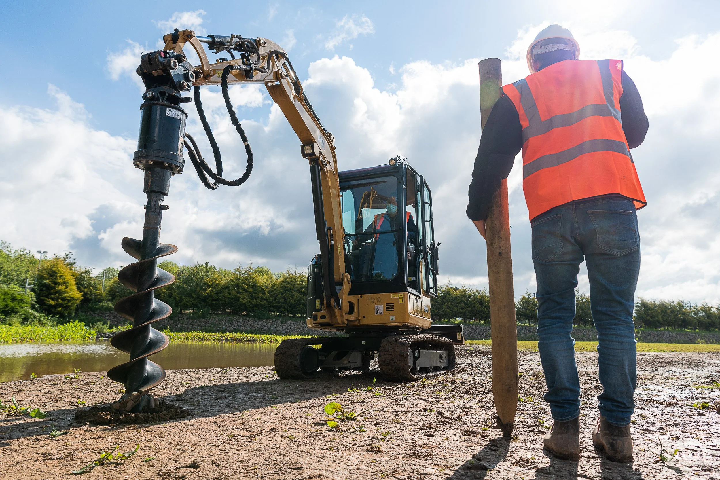 Worker guiding a post beside an excavator fitted with an auger.
