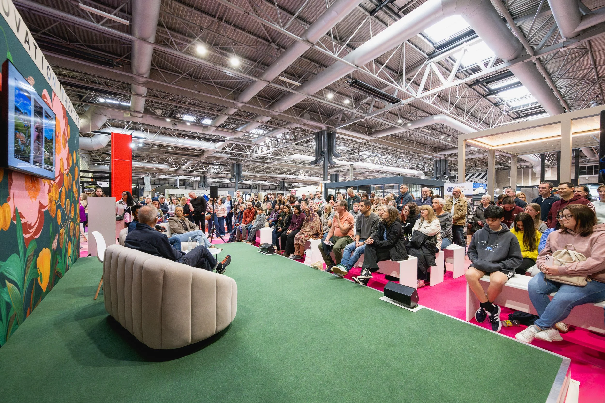 A seated audience watching a discussion in a busy Grand Designs Live presentation area.