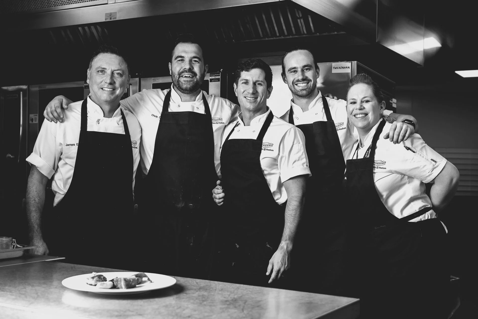 Black-and-white group portrait of five chefs standing together behind a kitchen counter with a plated dish in front.