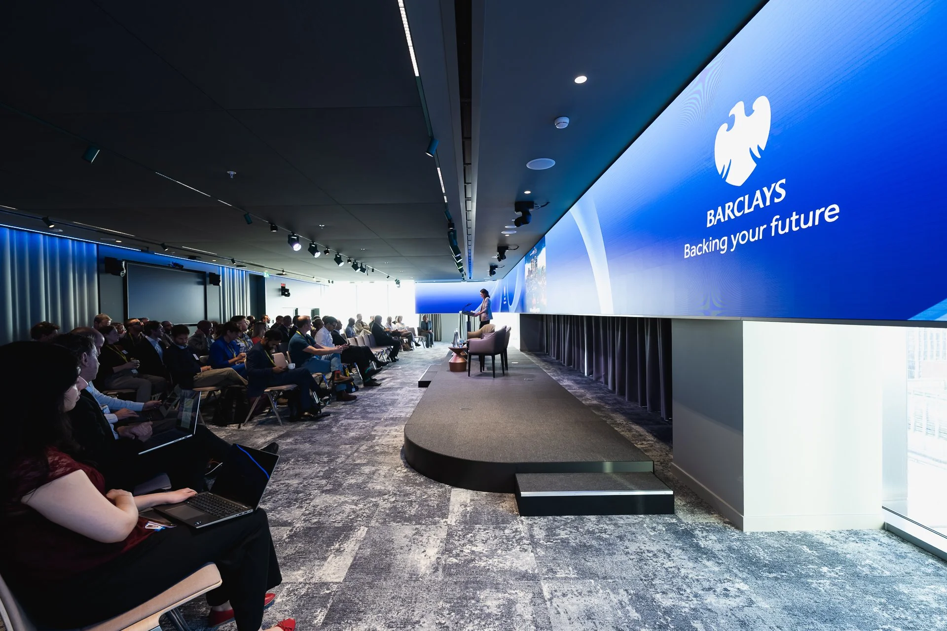 A wide view along the conference room, with seated delegates facing the stage and a large blue Barclays screen.