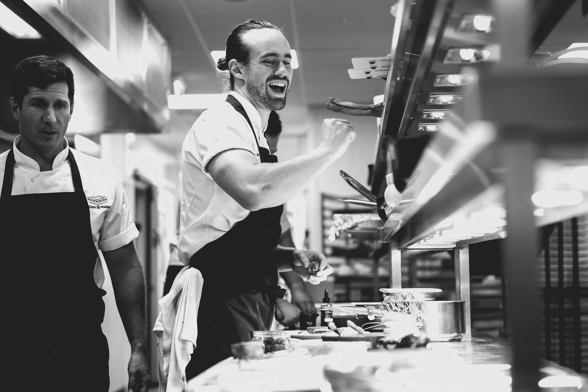 Black-and-white photograph of a smiling chef with one arm raised beside the kitchen pass, with another chef alongside.