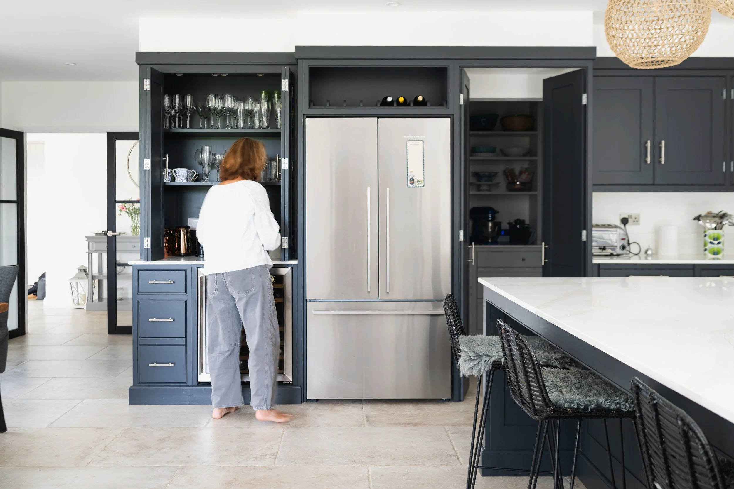 A woman standing at open kitchen cabinets beside a refrigerator and island.