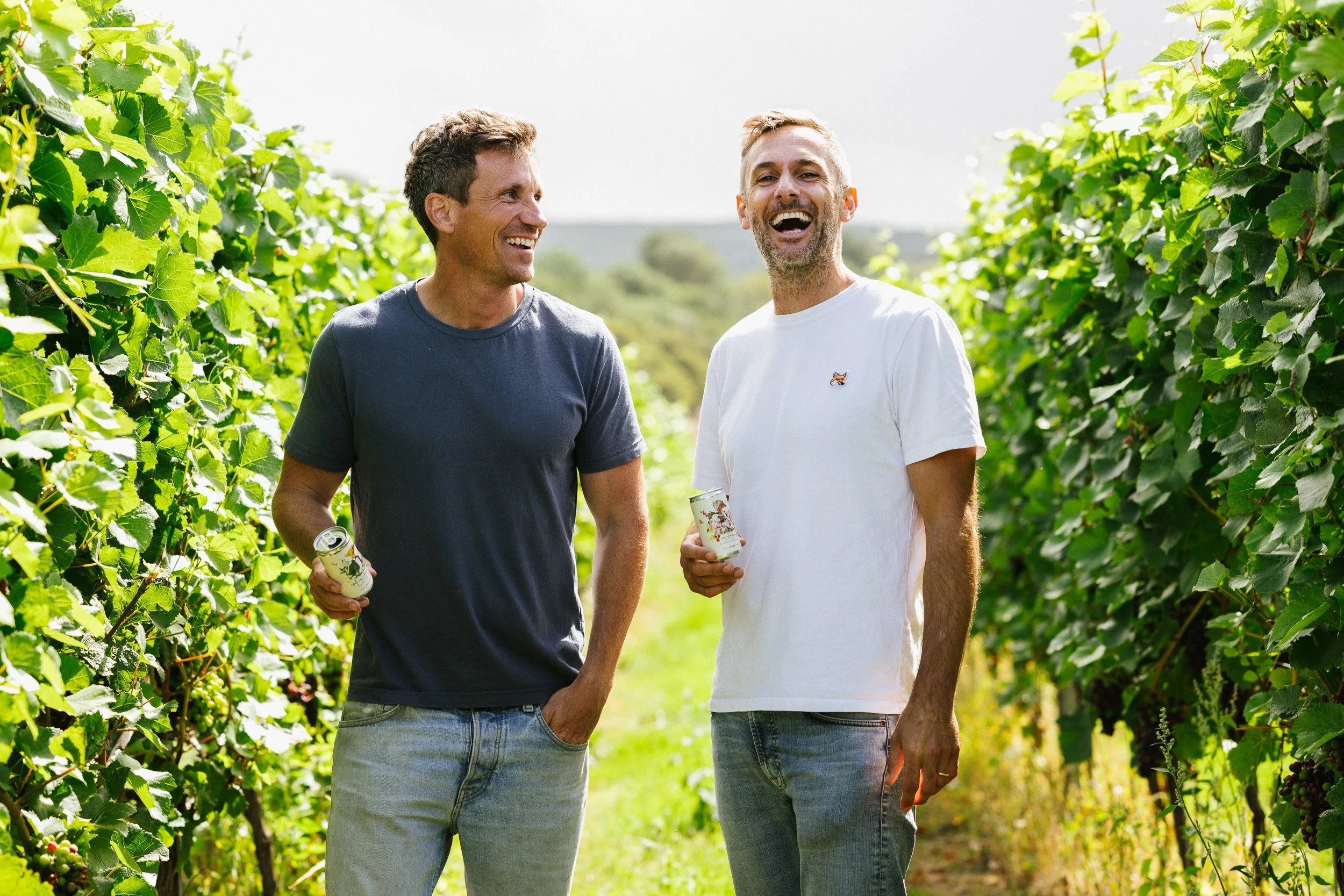 Two men laughing between vineyard rows while holding cans of wine.