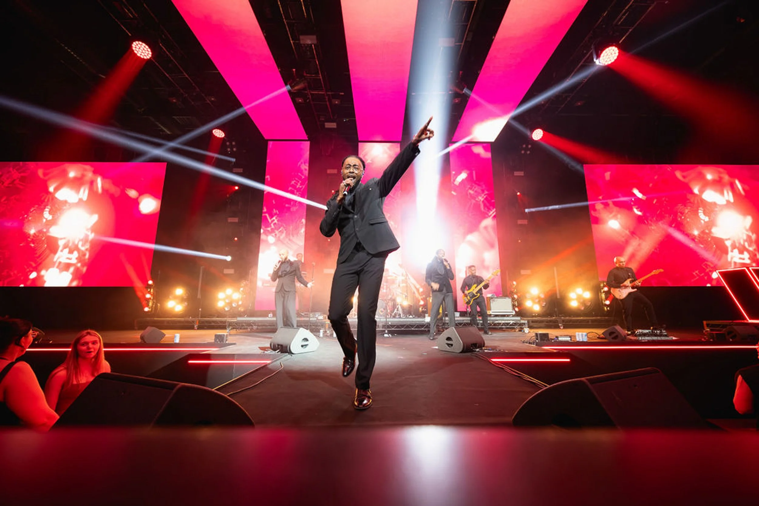 A singer performing at the front of the stage, with musicians and red lighting behind.