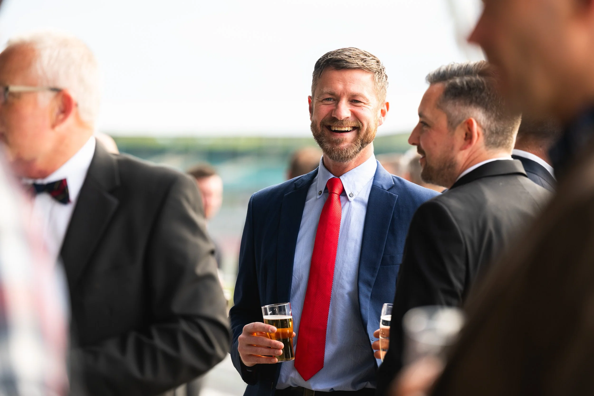 A smiling guest in a blue suit and red tie chatting with other guests over drinks.
