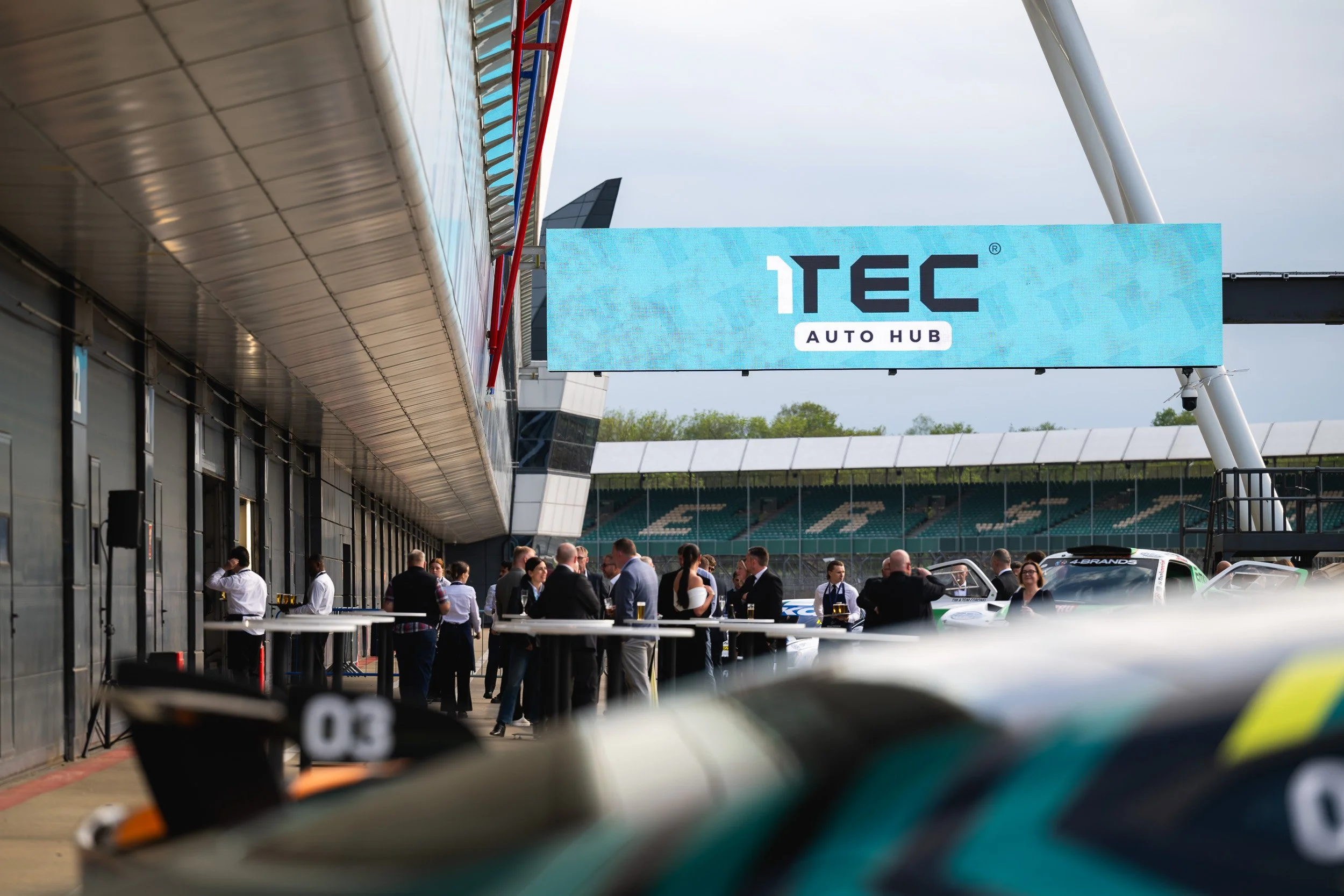 Guests gathering beside racing cars beneath a 1TEC Auto Hub sign at Silverstone.