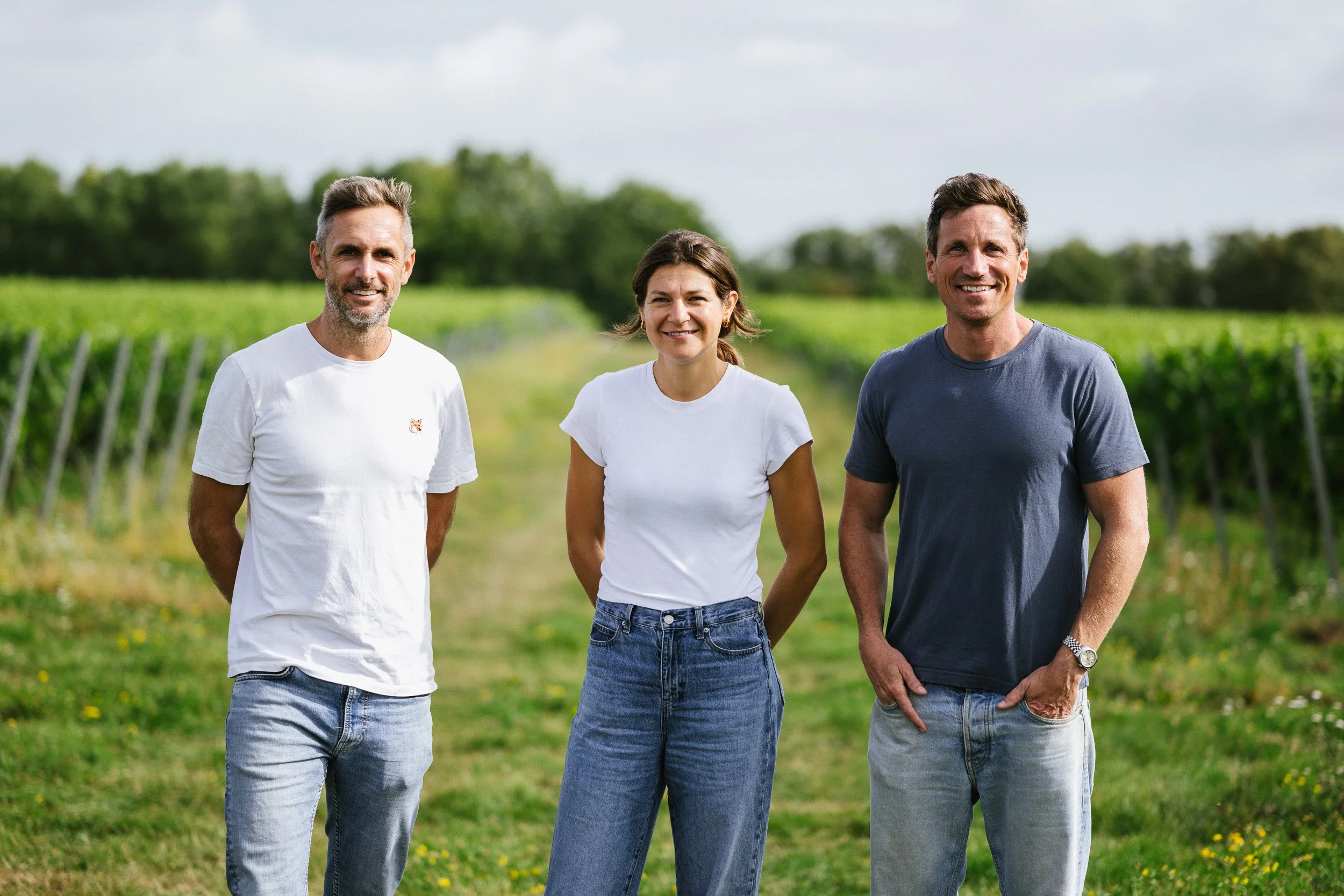 Three people standing together at a vineyard, with rows of vines behind them.