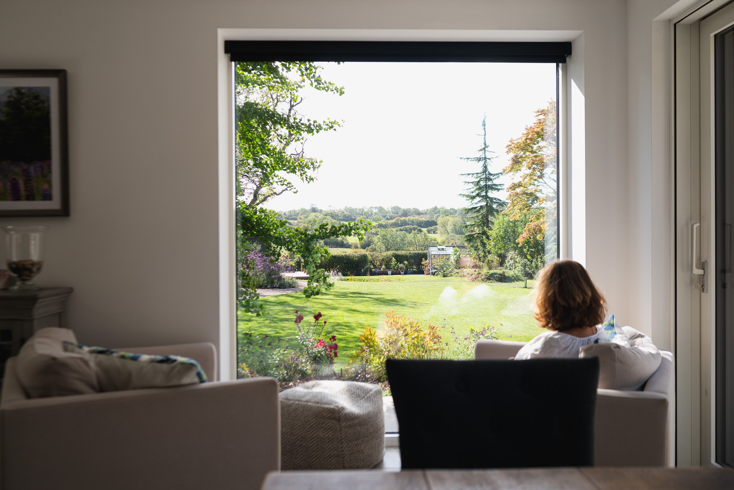 A woman seated beside a large window overlooking the lawn, trees and countryside.