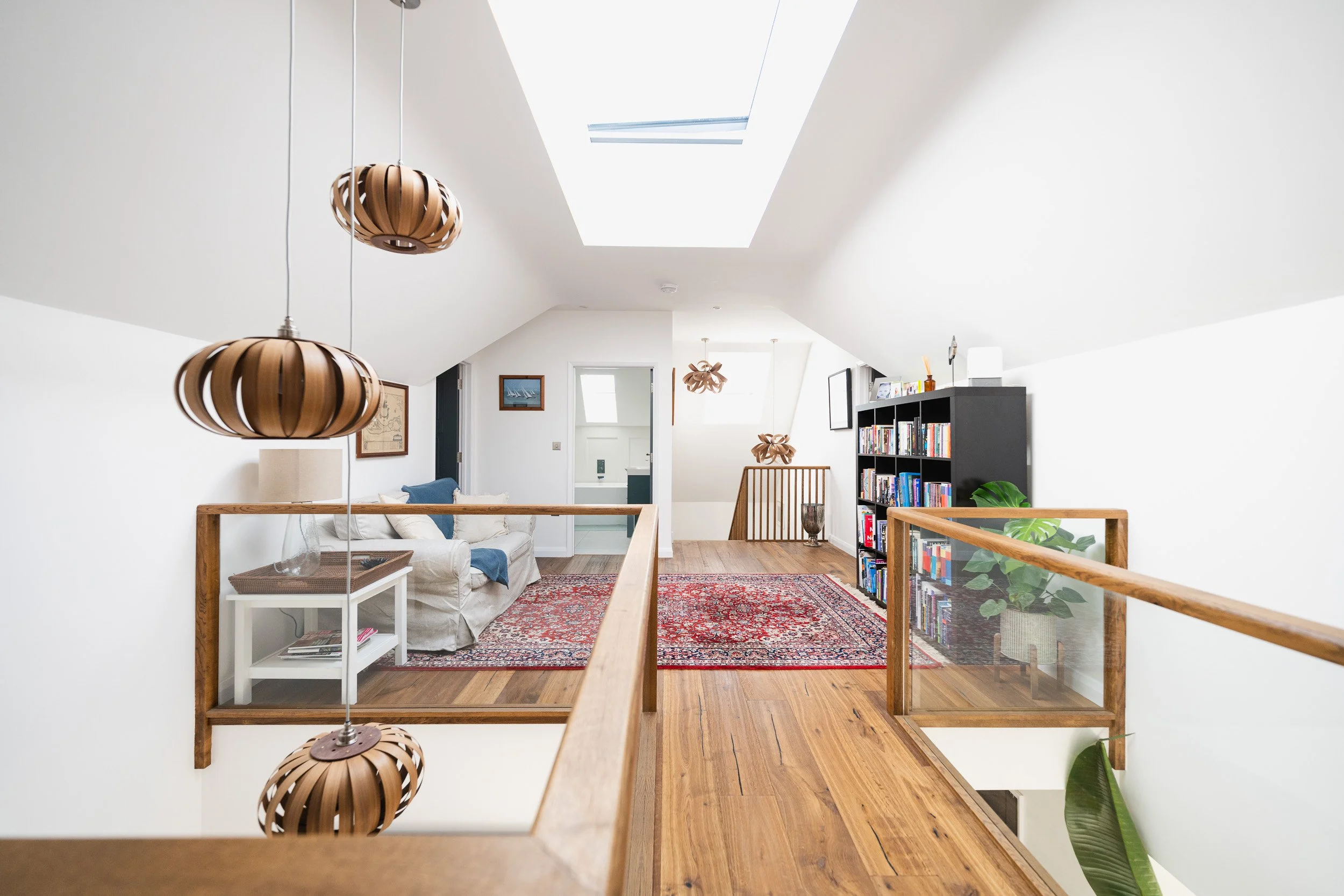 Timber-floored upper landing with glass balustrades, wooden pendant lights, a bookcase and seating beyond.