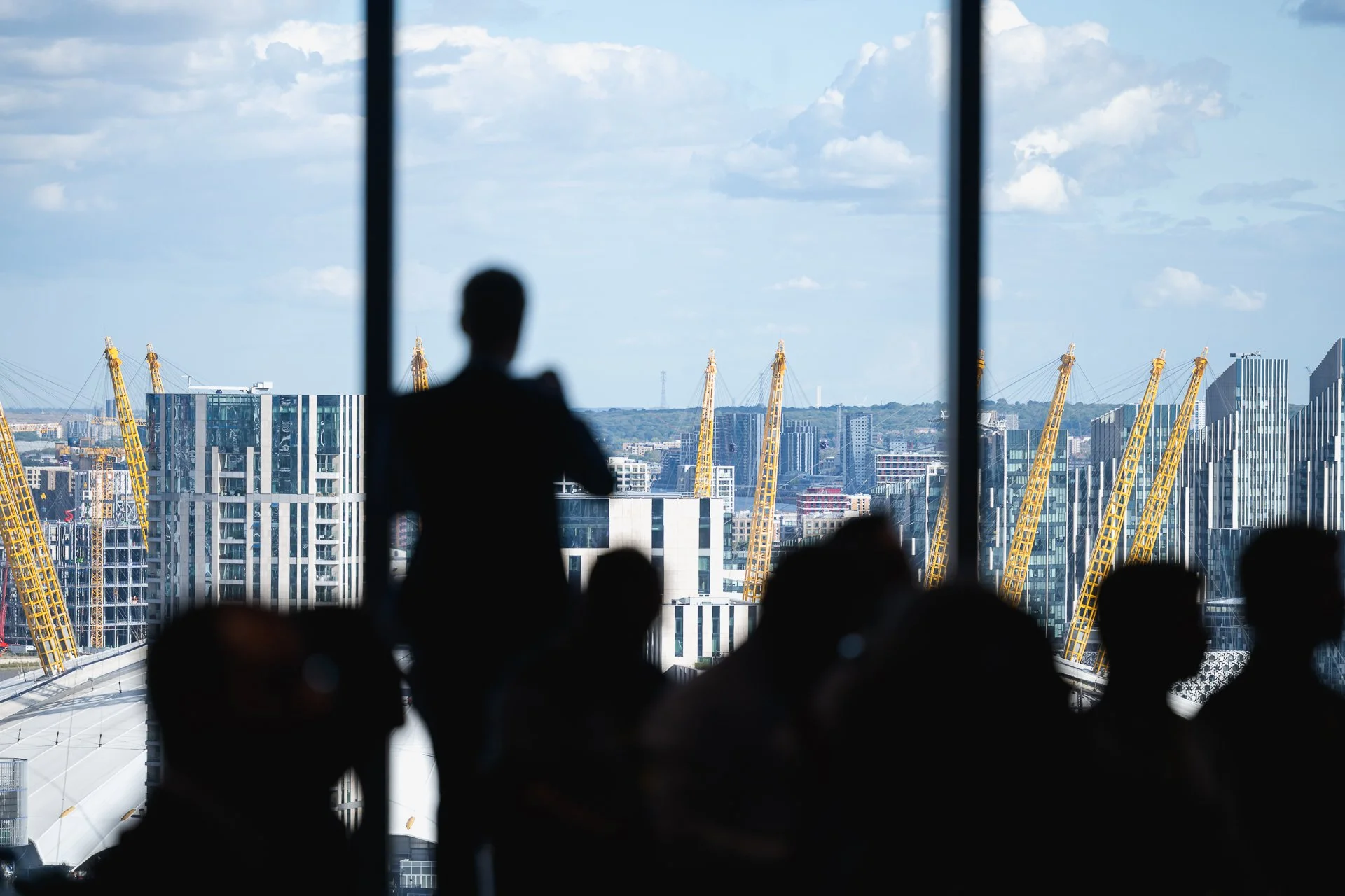 Delegates silhouetted against tall windows, with the yellow masts of The O2 and surrounding buildings outside.