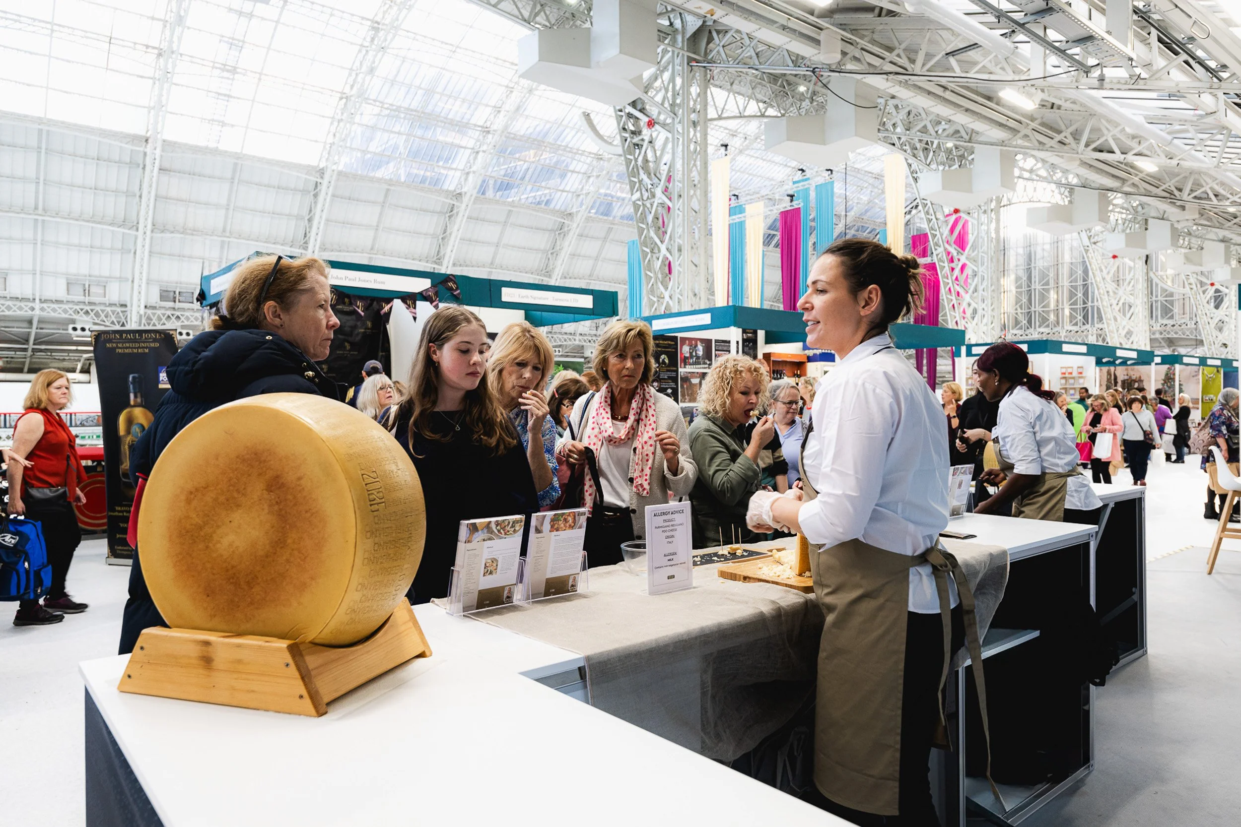 Visitors talking with an exhibitor beside a large cheese display at Olympia.