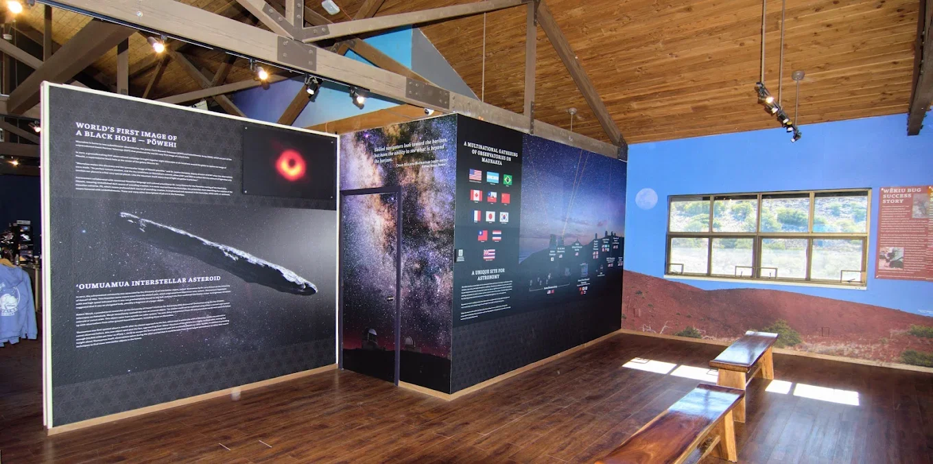 Interior Design work by Candice Kirby Designs for the Mauna Kea Visitors Center. An expansive exhibit wall detailing astronomical discoveries, including the first black hole image and 'Oumuamua, within a room featuring wooden benches and windows over