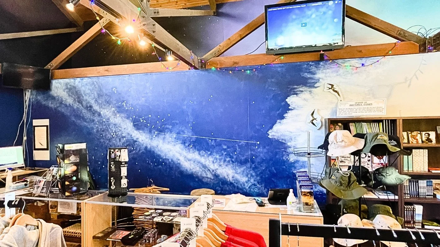 A retail area with a large blue mural of the Milky Way and constellations behind a wooden sales counter, shelves with merchandise like hats and books, string lights, and another television. Candice Kirby Designs' work on the Mauna Kea Visitors Center