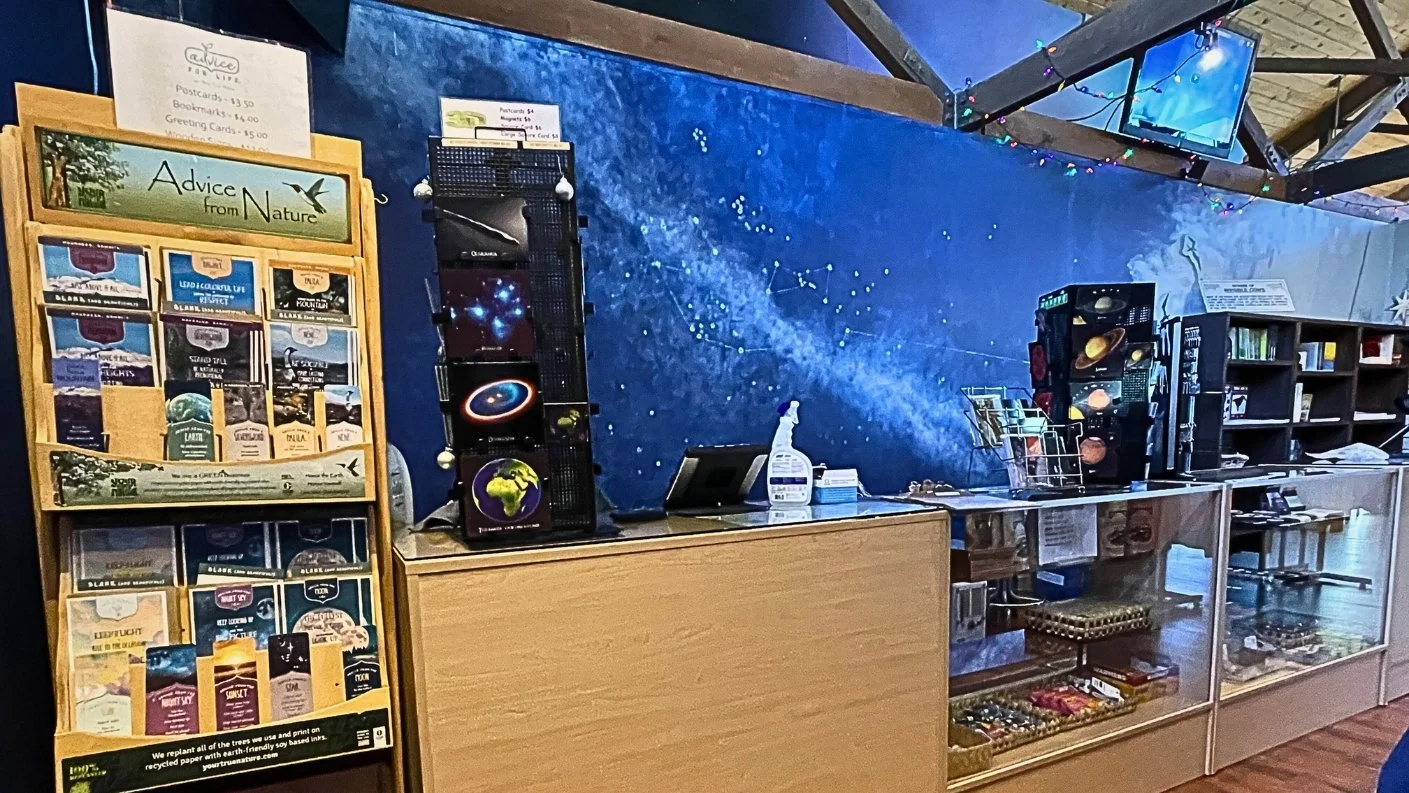 Candice Kirby Designs' interior work for the Mauna Kea Visitors Center. A retail section with a prominent blue mural of the Milky Way and constellations, a wooden sales counter, and shelving units stocked with various merchandise like hats, books, an