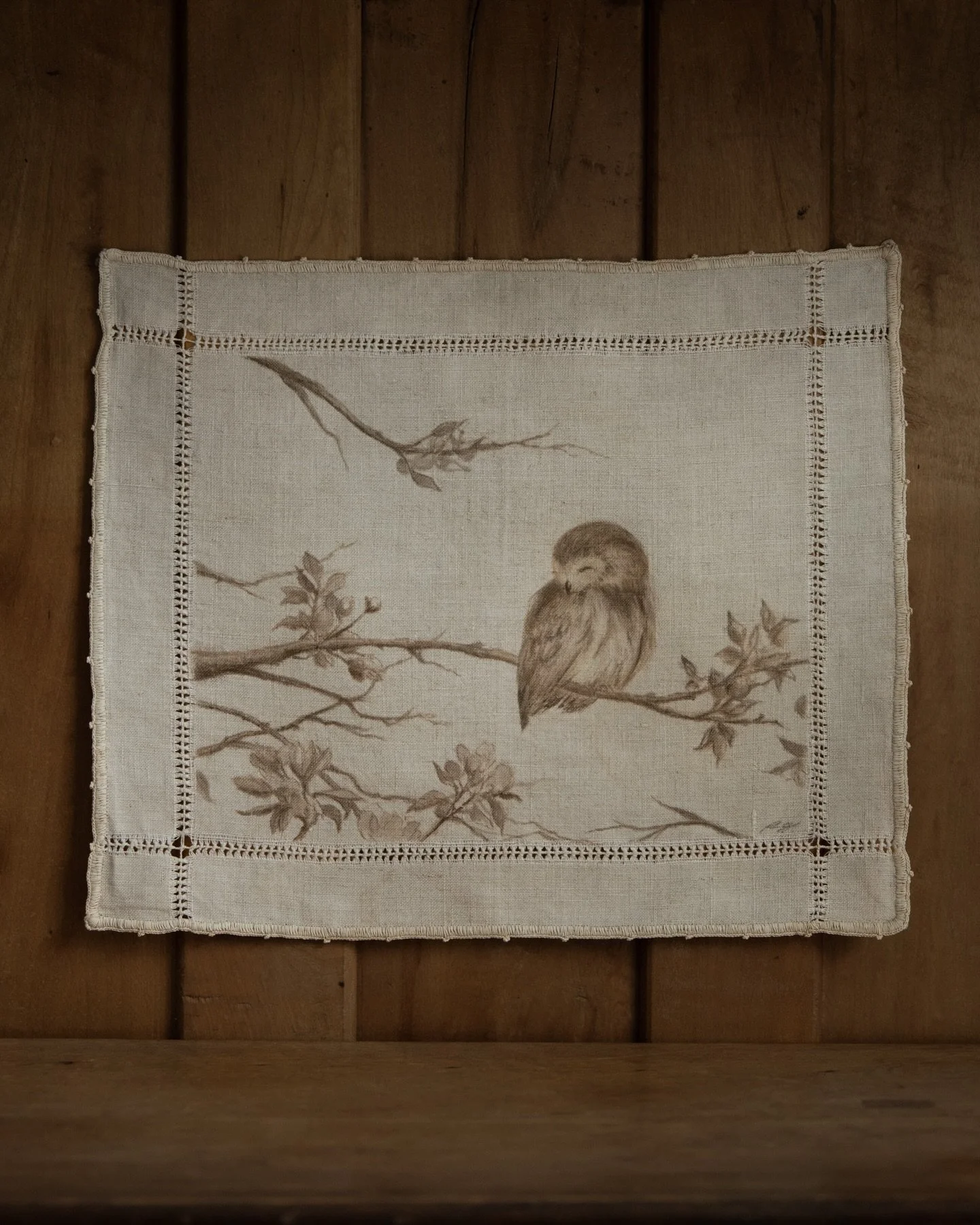 A northern saw-whet owl resting on a blossoming branch. The age of the hemstitched linen naturally slows the scene down. Once part of a domestic setting, it brings a lived-in softness into the painting, gently diffusing everything.