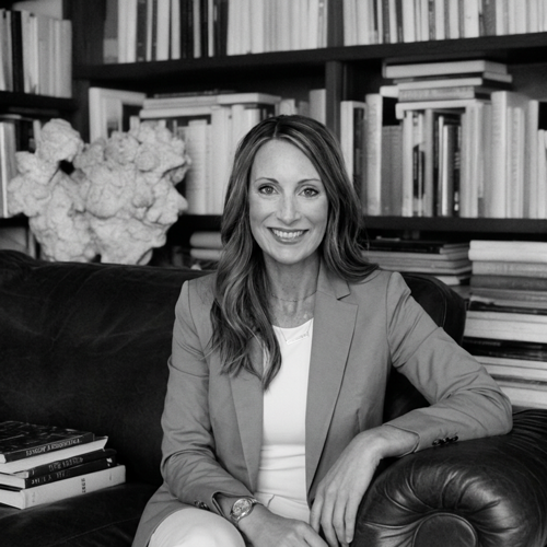 A woman with long hair smiling, sitting on a black leather couch in front of a bookshelf filled with books.