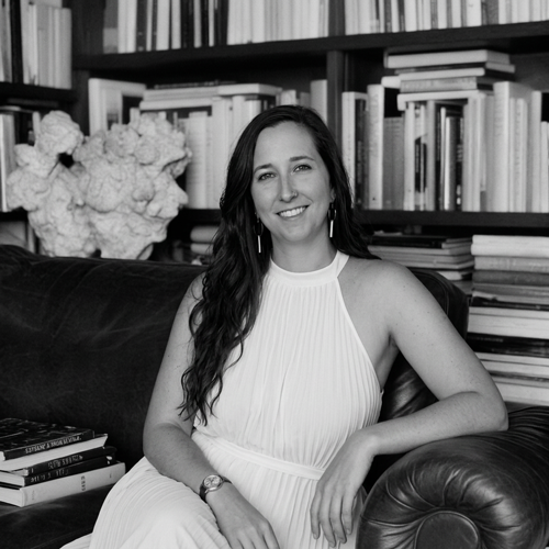 A woman with long wavy hair in a sleeveless white dress sitting on a couch in front of a bookshelf filled with books.