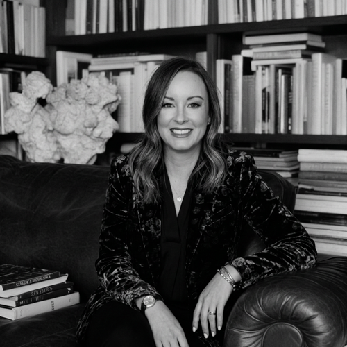 A smiling woman sitting on a leather sofa in front of a bookshelf with flowers and books.