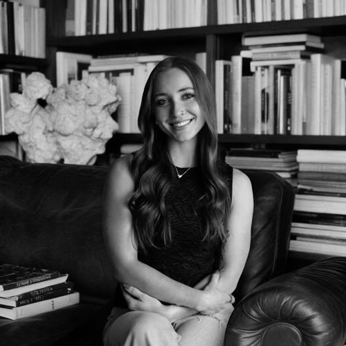 A young woman with long wavy hair smiling and sitting on a couch in a room with bookshelves filled with books behind her.