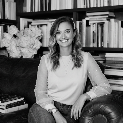 A woman with wavy hair, wearing a light-colored blouse with sheer sleeves, sitting on a black leather couch in a room with bookshelves and a large floral arrangement in the background.