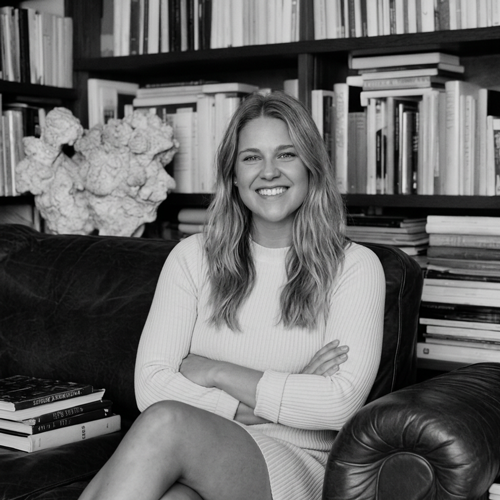 A woman with long wavy hair sitting on a black leather couch in front of a bookshelf filled with books, smiling at the camera.