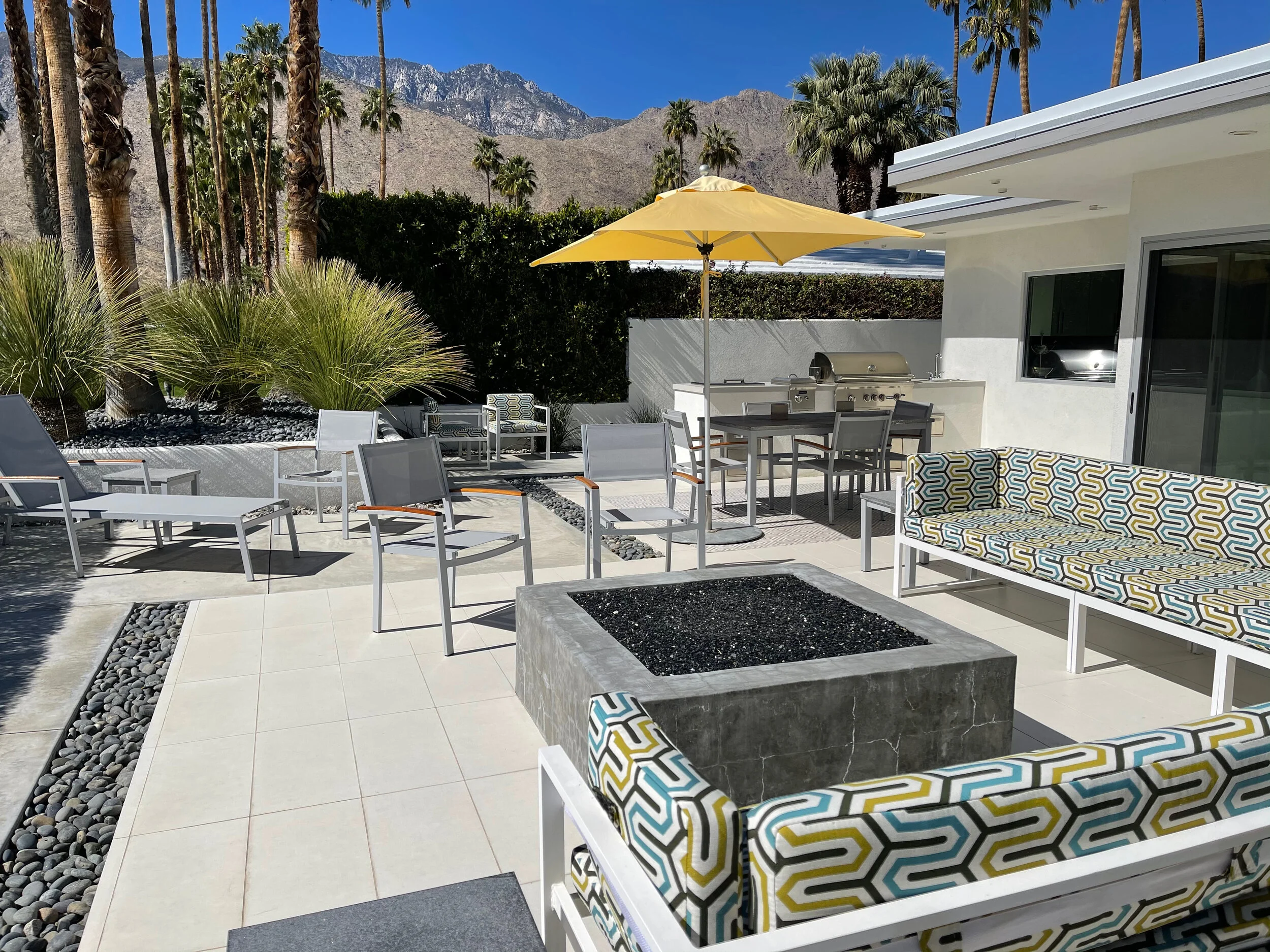 Modern architectural home with resort-like patio and fully-equipped outdoor kitchen and outdoor dining set, custom upholstered patio lounge chairs in bright mid-century geometric pattern Sunbrella fabric, glass rock concrete built firepit for indoor