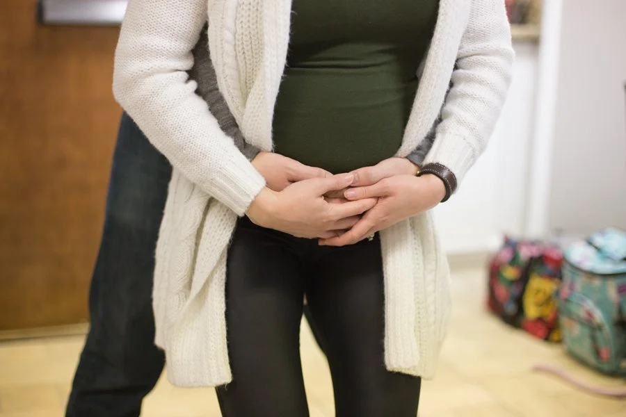 A woman holding her pregnant belly with both hands indoors, with bags visible in the background.