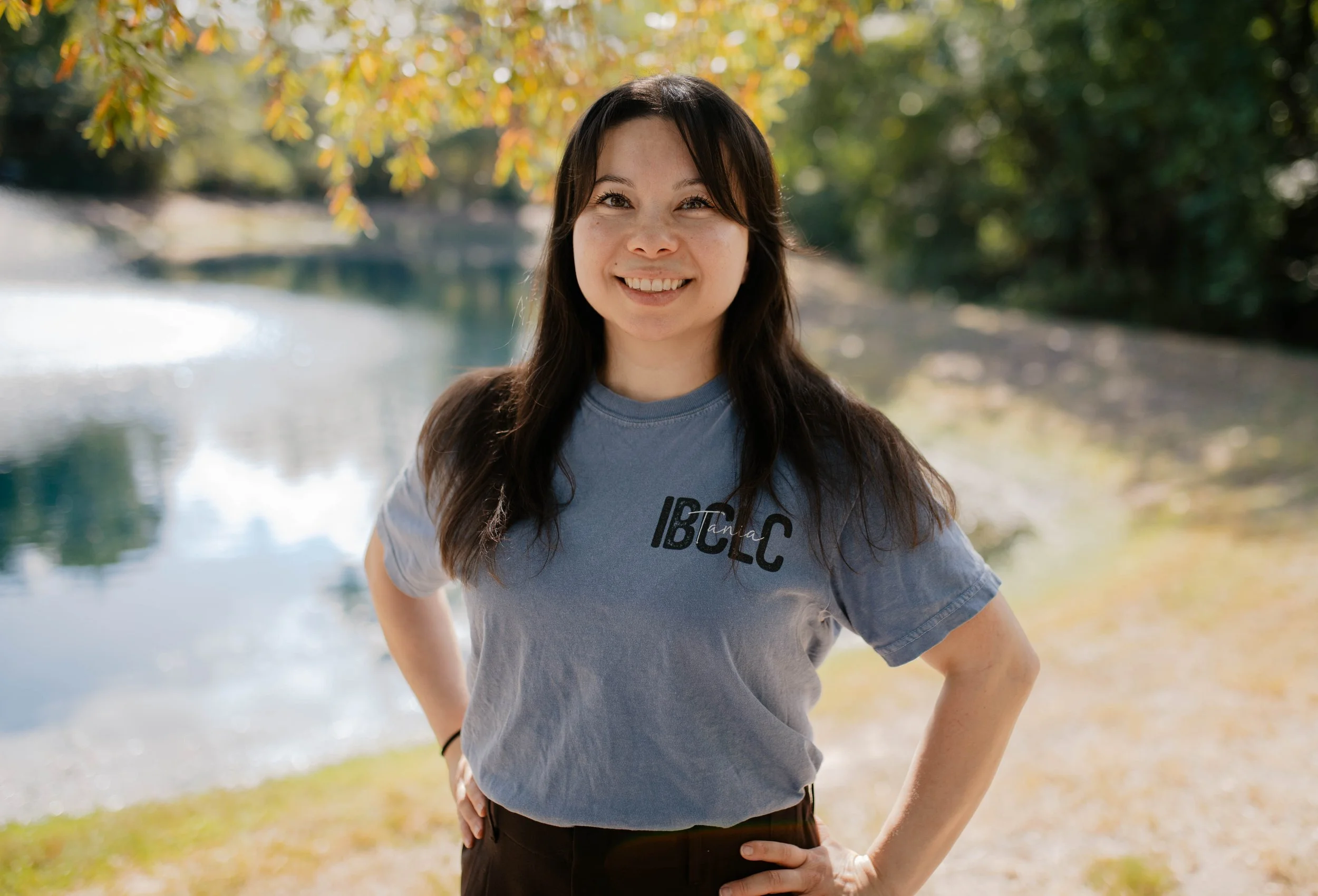 Tania Nuezca, IBCLC and The Bradley Method childbirth educator posing for a headshot. She is outdoors near a body of water surrounded by trees with autumn leaves.