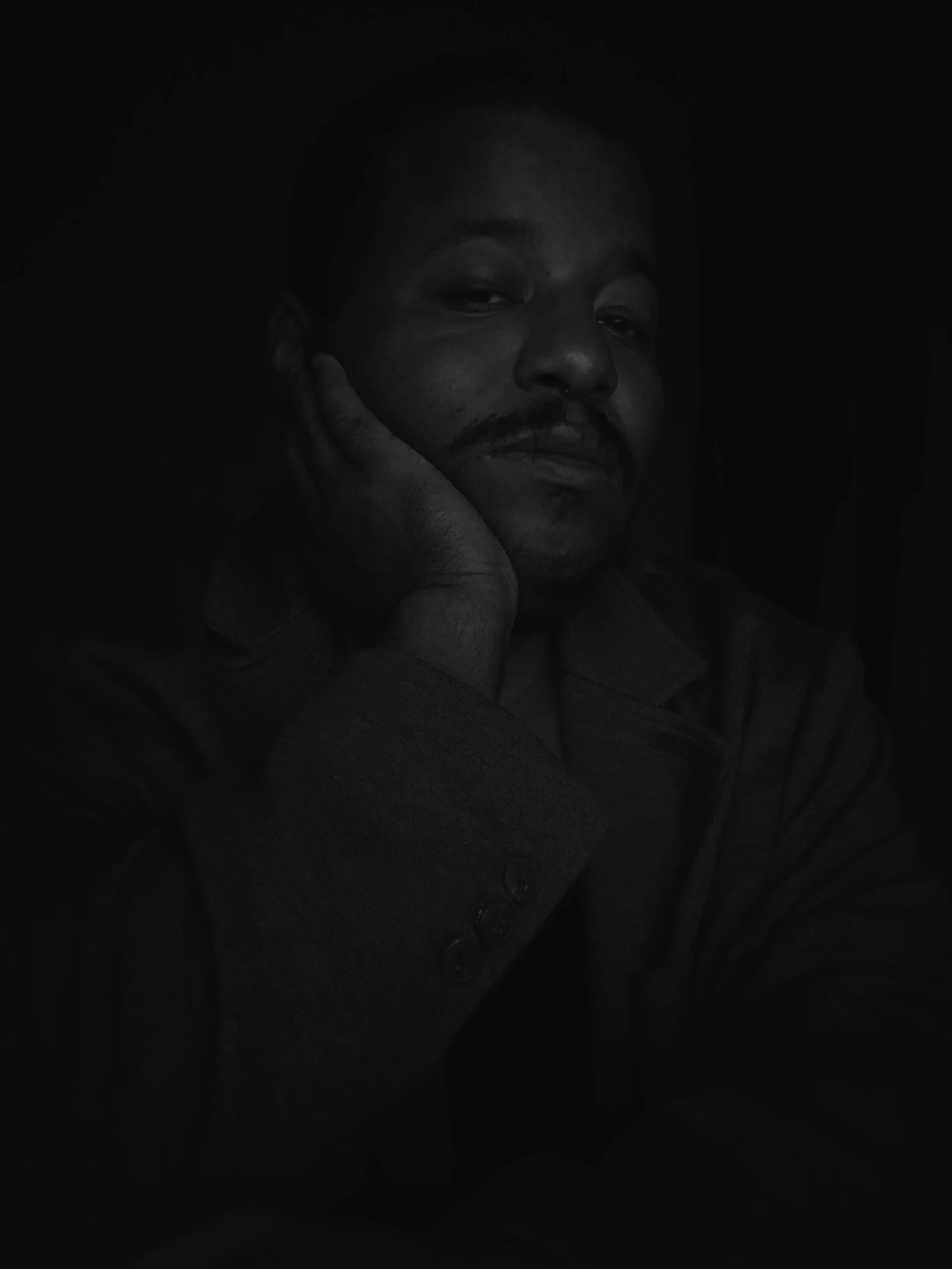 A black and white photograph of a man with facial hair, resting his hand on his face, with a serious expression, wearing a coat with buttons.