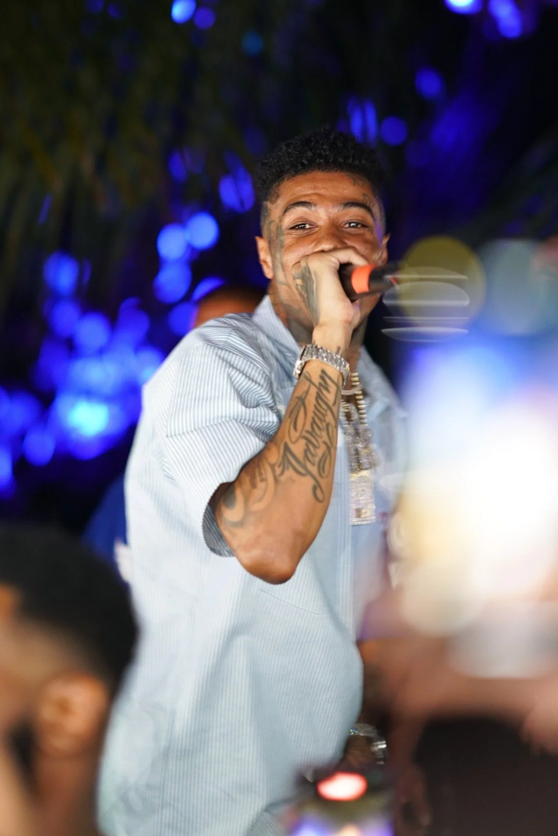 A man with tattoos holding a microphone, smiling, and singing or speaking at a party or event, with people taking photos in the foreground and colorful blue lights in the background.