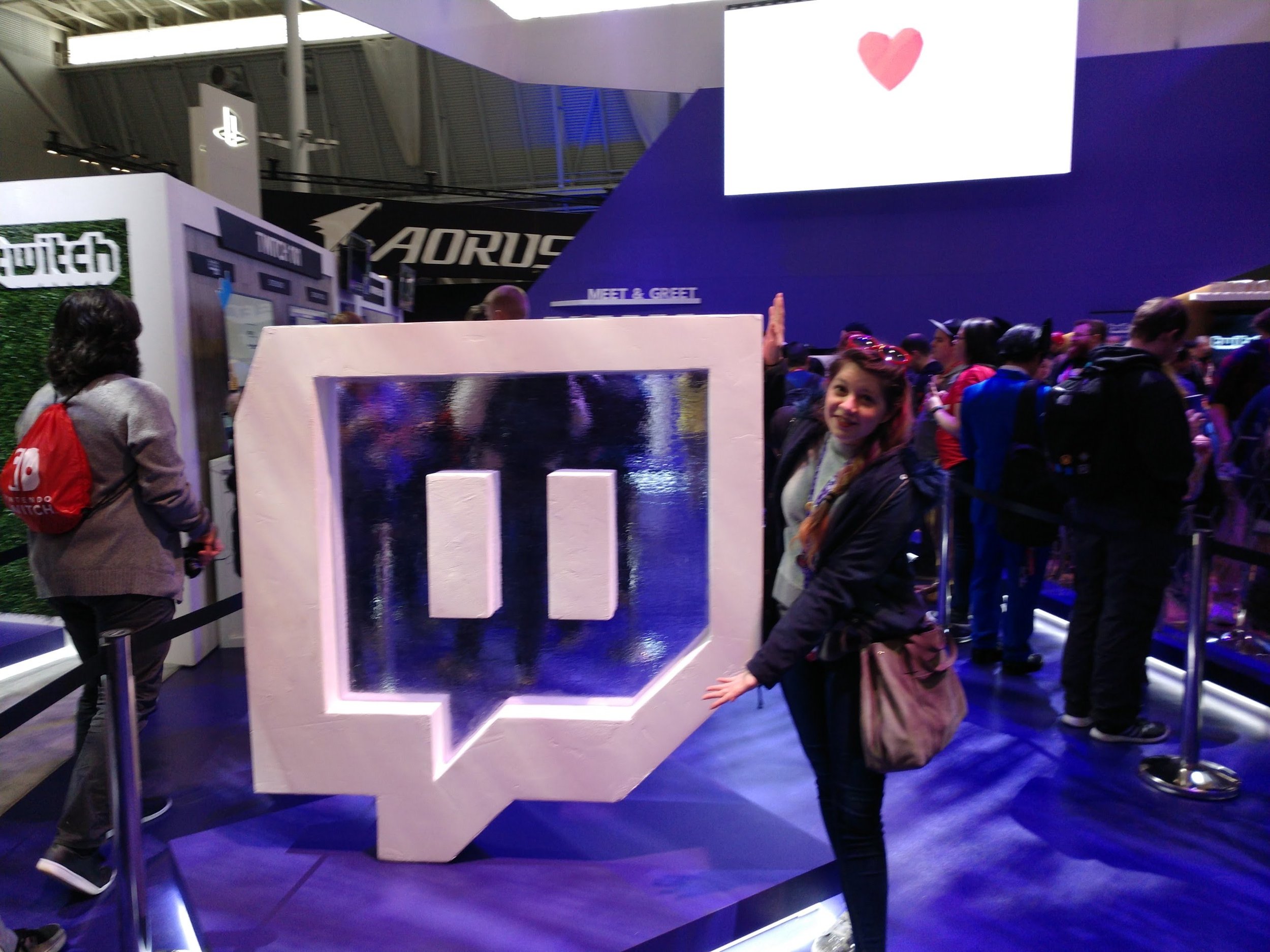 A woman at a Twitch booth with a large Twitch logo in a speech bubble shape, in a crowded event space, with a small heart symbol shown on a screen above.