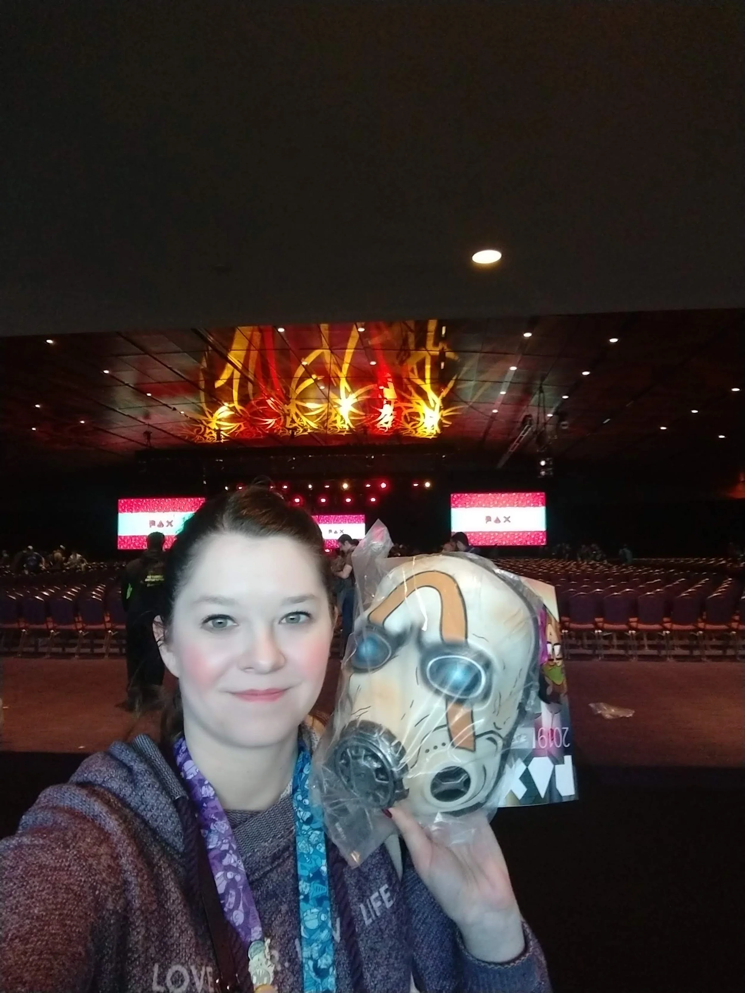 A woman takes a selfie at a large gaming conference or event hall, holding a mask resembling one from a video game in front of her face. The hall is dimly lit with rows of empty chairs and a big stage with screens in the background.