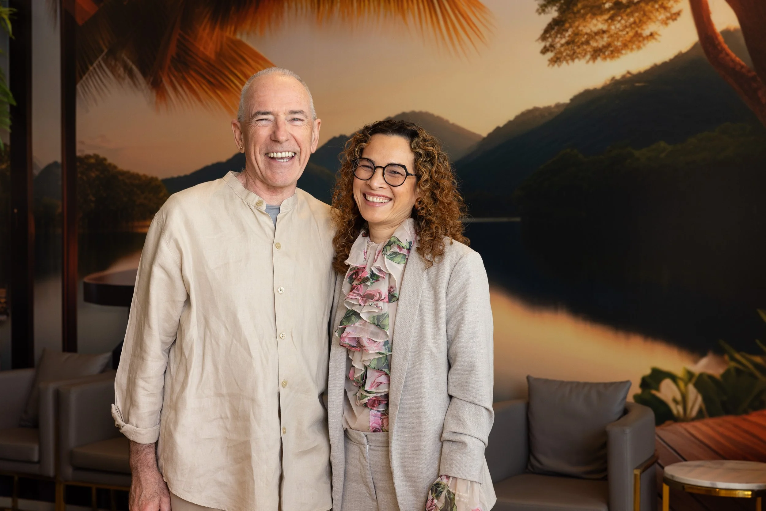 A man and woman smiling and standing together indoors with a large scenic mural of a lakeside and mountains at sunset on the wall behind them.