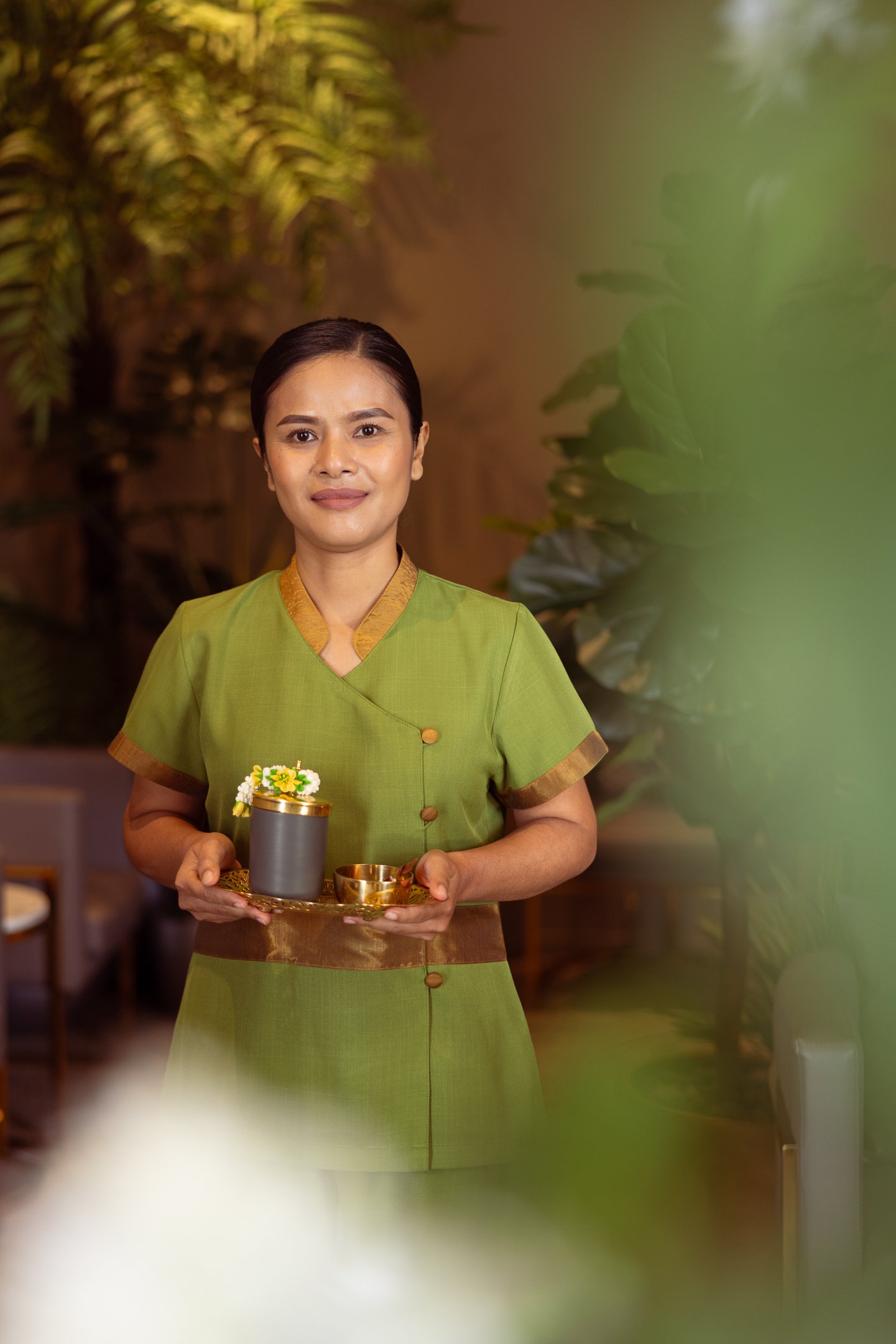 Thai Touch's Praewa, holding a tray with a gray container, a small bowl, and yellow flowers, standing in a lush indoor setting.