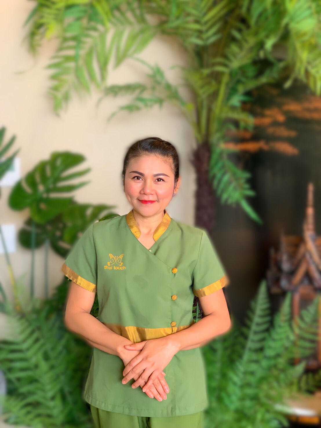 Thai Touch Massage Therapist, Mint, standing in front of a tropical-themed background with large green plants.