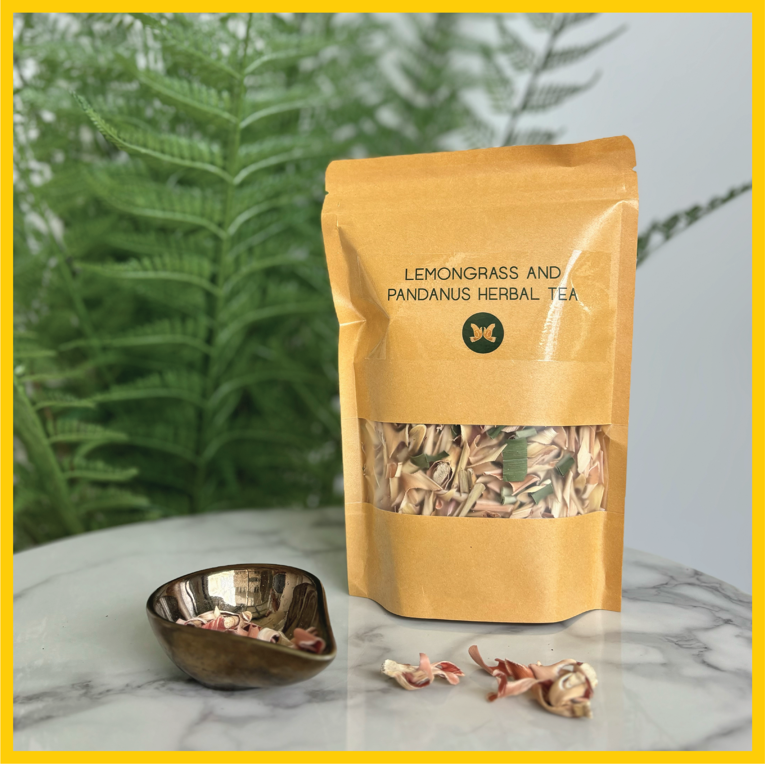 A brown paper bag labeled 'Lemongrass and Pandanus Herbal Tea' with a transparent window showing dried herbs, placed on a marble surface with some herbs spilled nearby, against a background of green fern leaves.