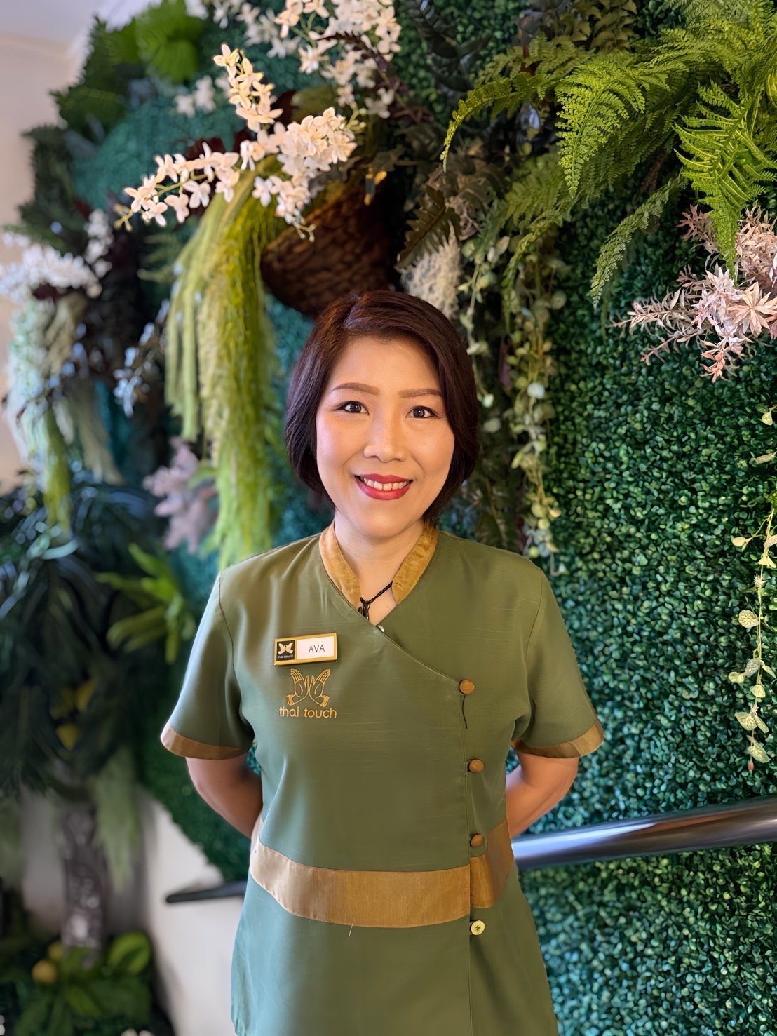 Thai Touch Massage Therapist Ava, standing in front of a lush green floral wall with pink and white flowers.